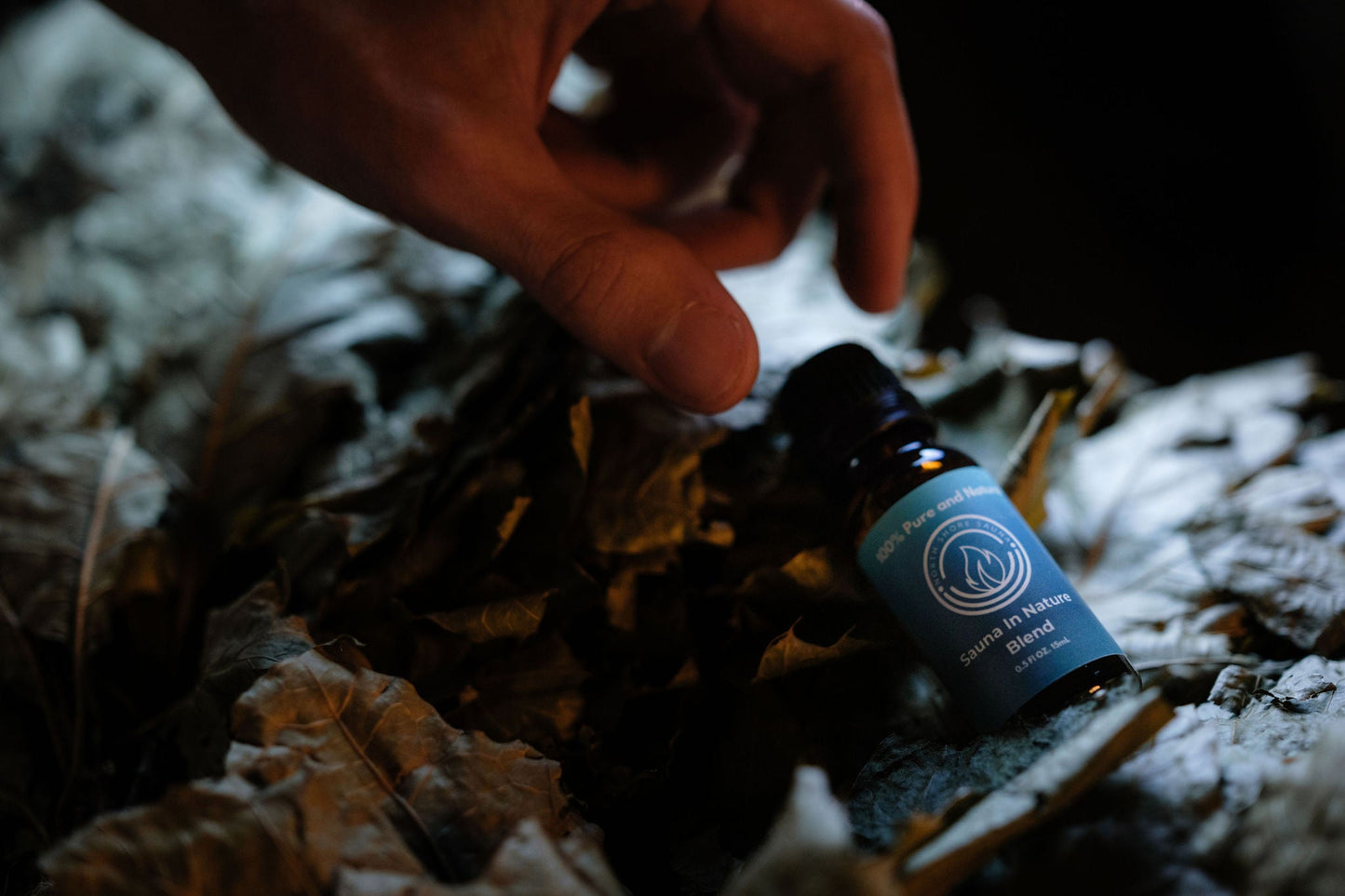 Essential Oil Blend | Sauna In Nature bottle on a bed of leaves in a dimly lit sauna setting.