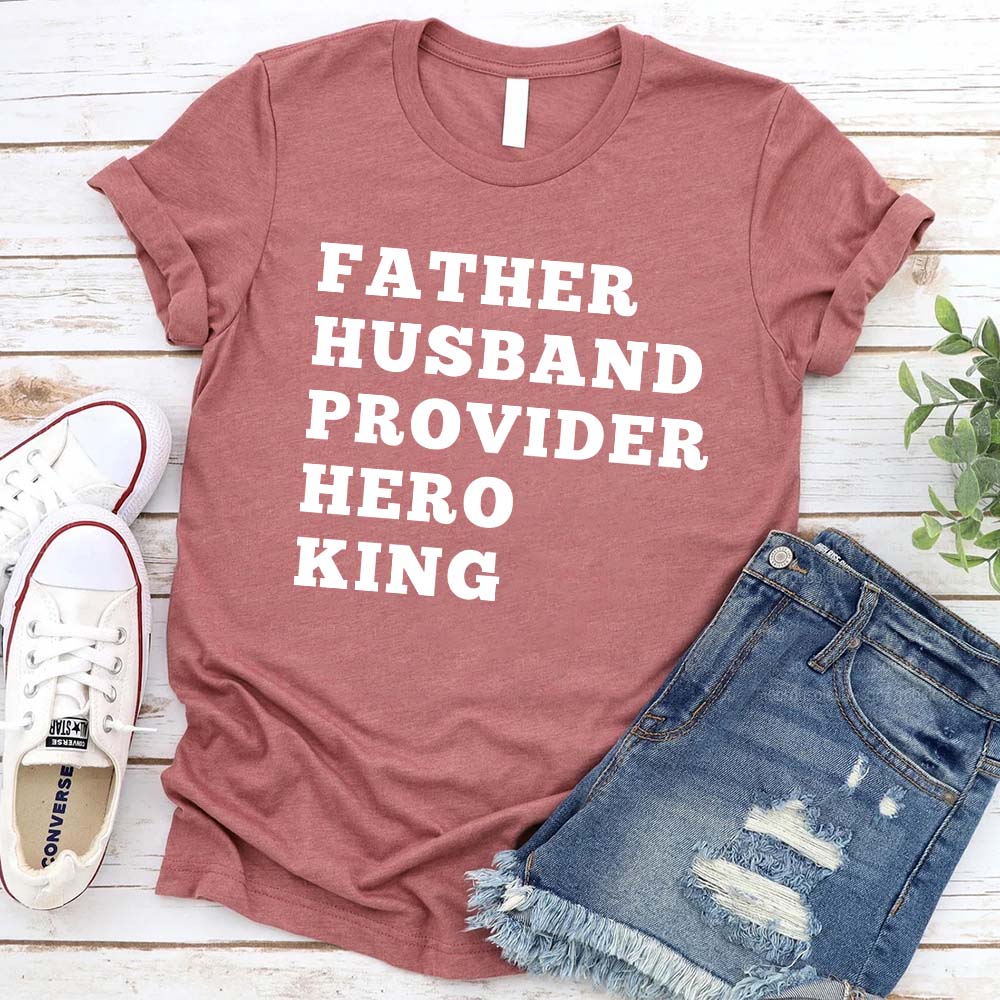 Alt Tag: Father Husband Provider Hero King T-shirt in red with text, laid next to denim shorts and white sneakers.