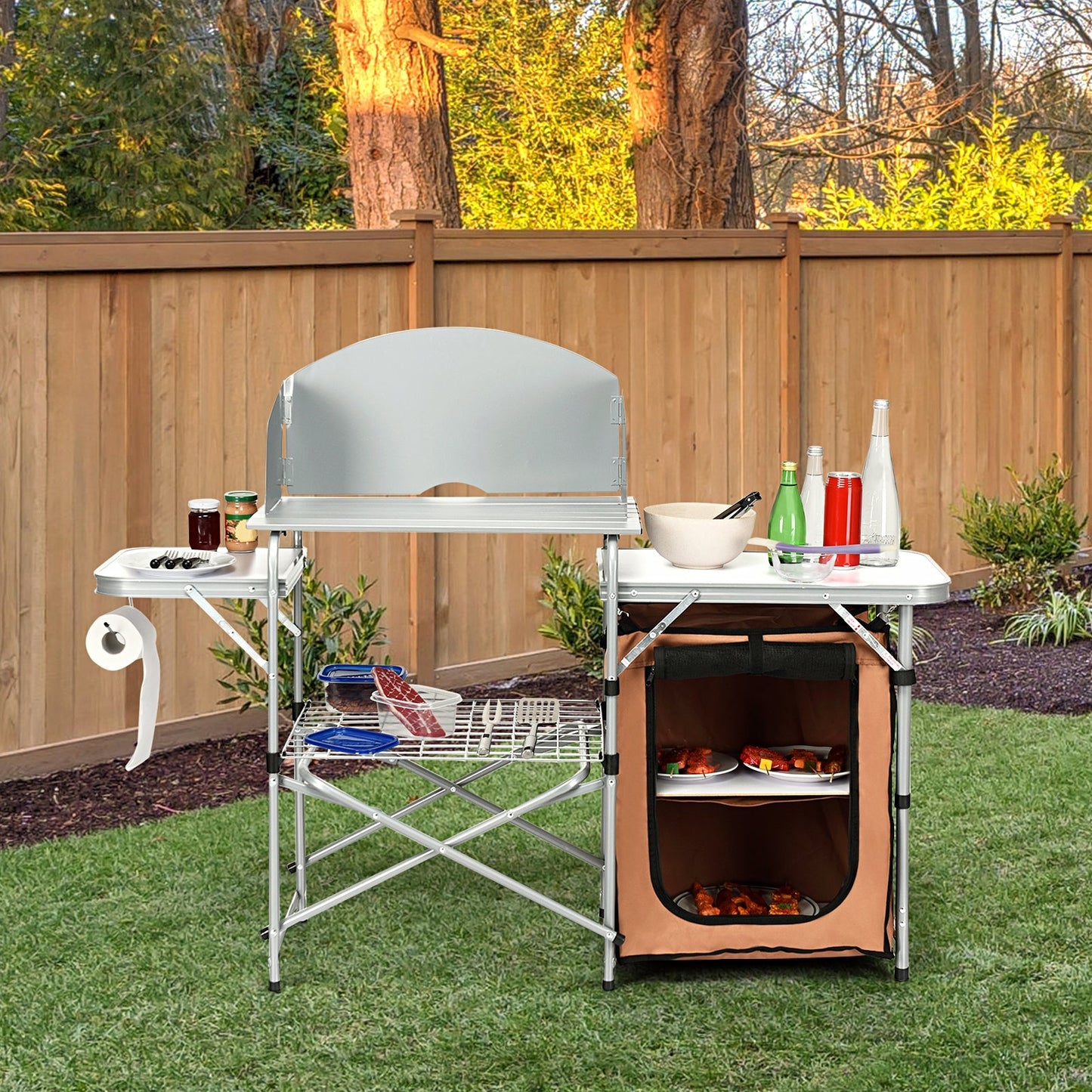 Foldable Outdoor BBQ Portable Grilling Table With Windscreen Bag in a garden setting, featuring side tables and storage compartments.