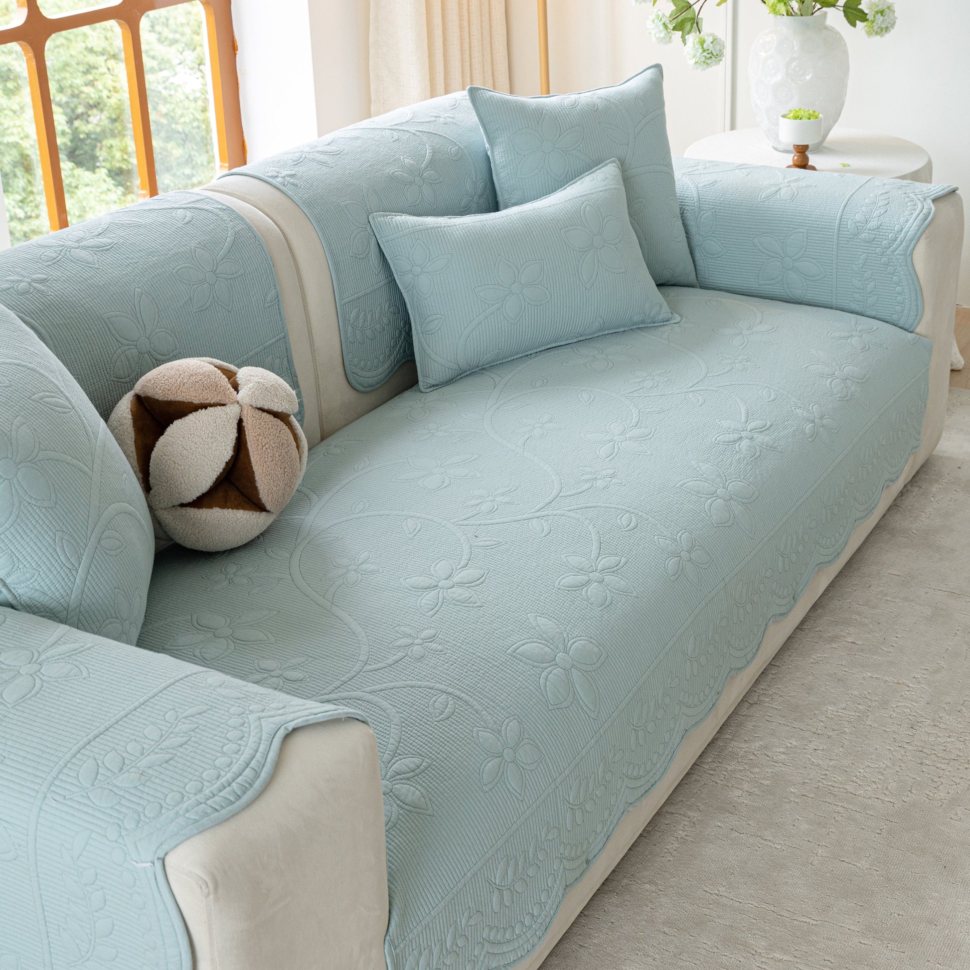 Floral Cotton Quilted Sofa Cover, Non-Slip Couch Protector with Scalloped Edges on a light blue sofa in a bright living room.