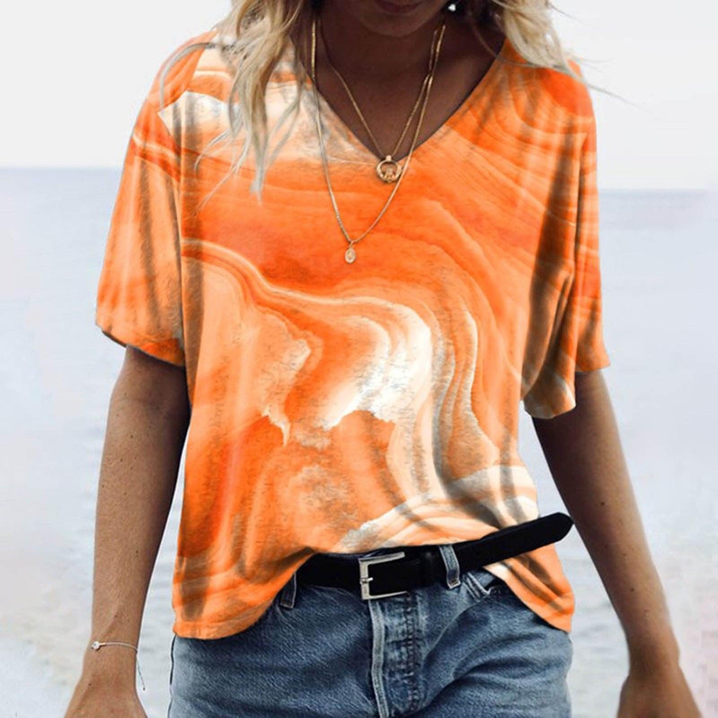 Woman wearing Women's Spring Glitzy V-Neck Short Sleeved Marble Print Top in vibrant orange marble design on the beach.