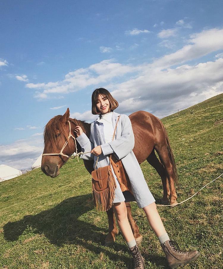 Woman in coat holding Dakota Tasseled Boho Leather Bag - Trendy Large Tassel Shoulder Bag with horse in open grassy field