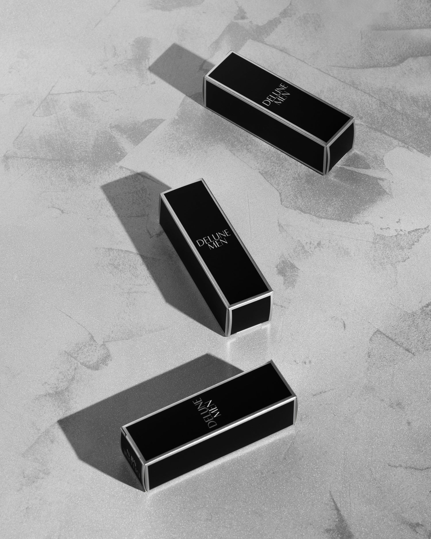 Three rectangular black lipstick tubes on a textured surface, casting dynamic shadows in a black-and-white aesthetic.