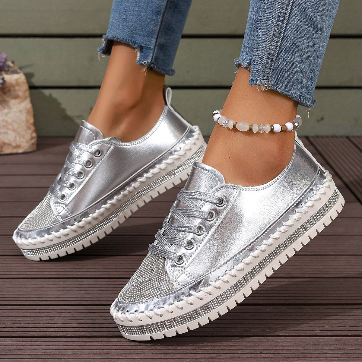 Metallic silver sneakers with a chunky sole on a wooden deck, worn with frayed hem jeans and a beaded anklet.