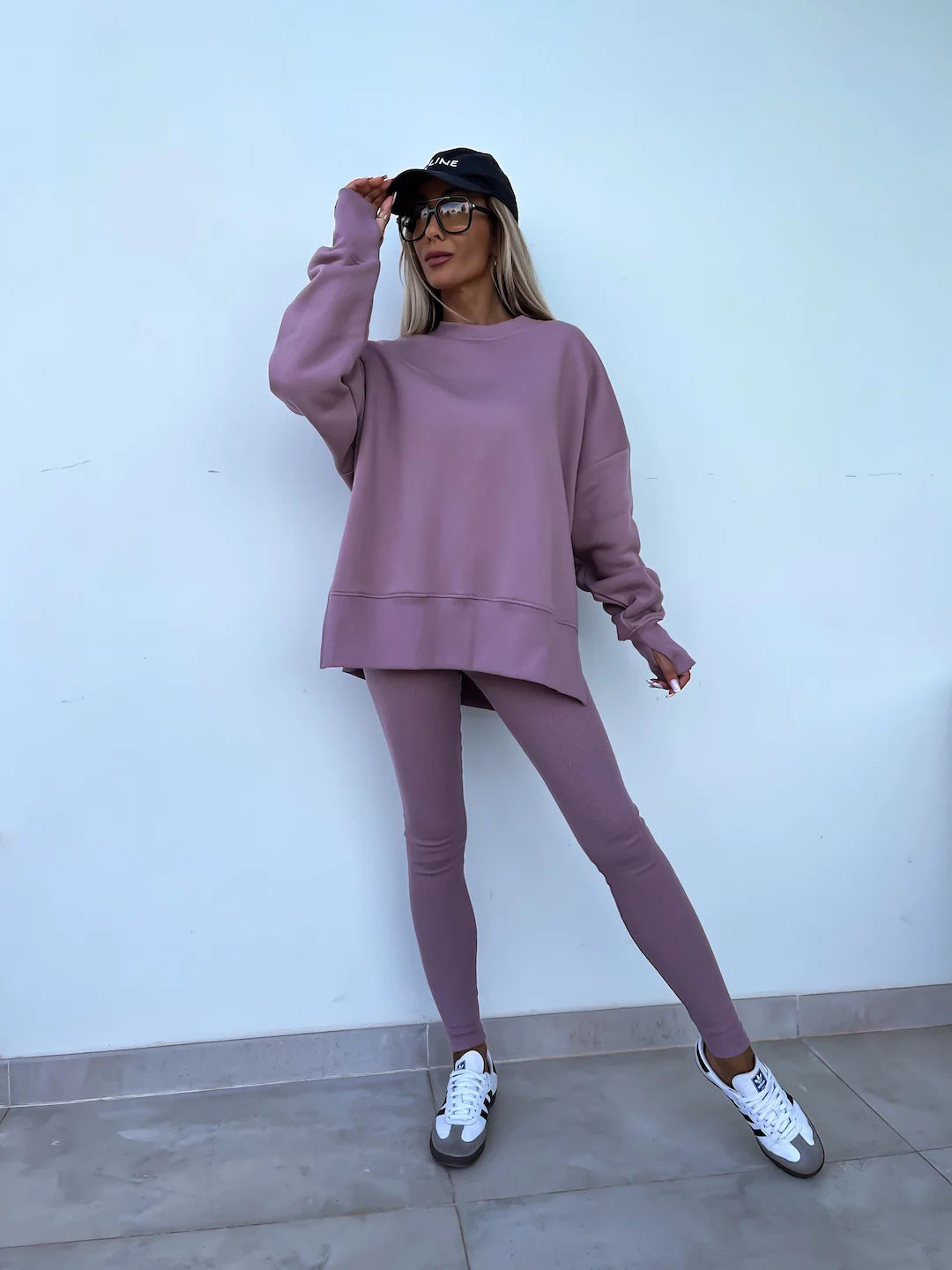 Woman wearing Women’s Casual Sweater Suit – Loose Split Top & Fitted Trousers Set in mauve, standing against a white wall with a cap and sneakers.