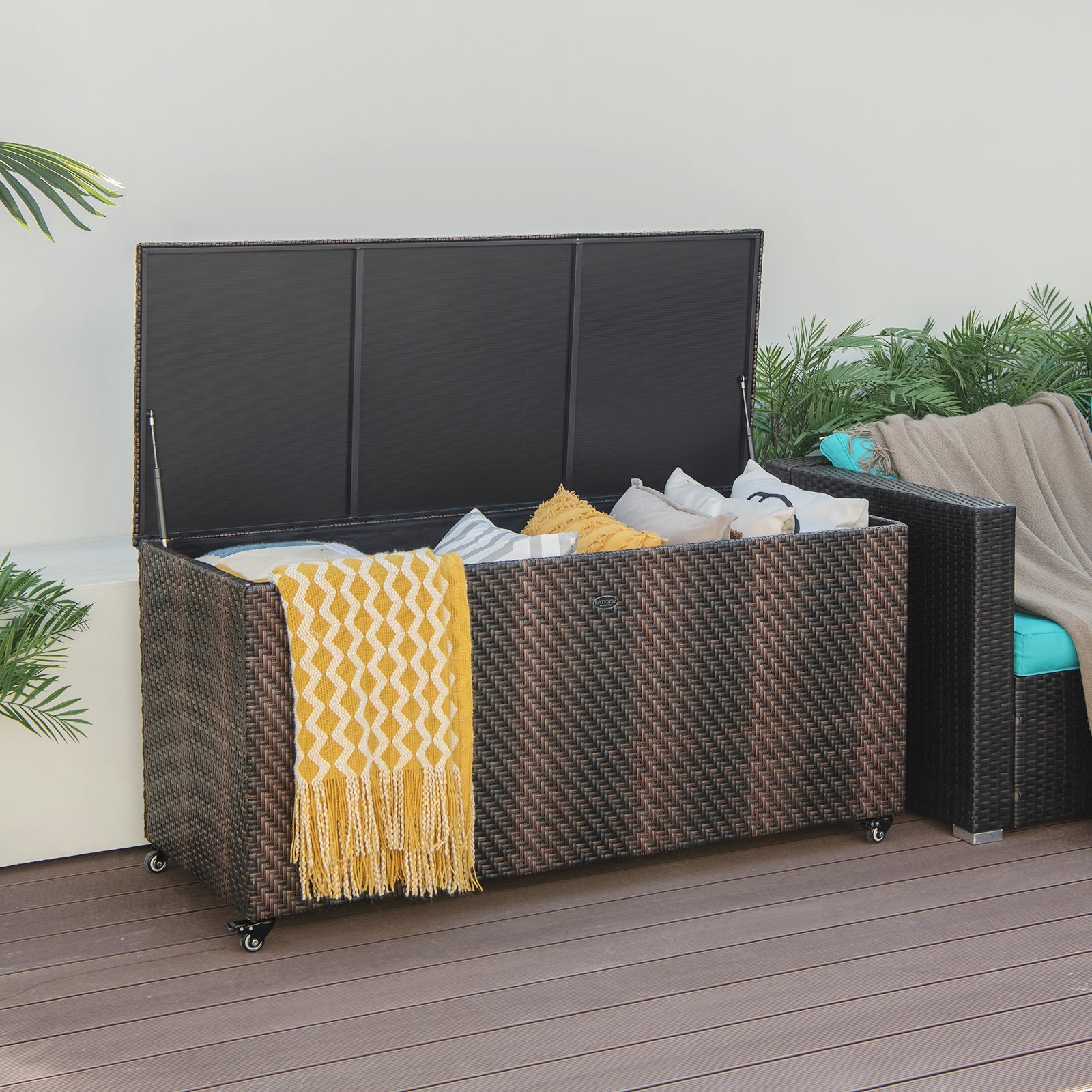 96 Gallon PE Wicker Outdoor Storage Box with 4 Wheels-Brown, open with cushions and a yellow blanket, on a patio.