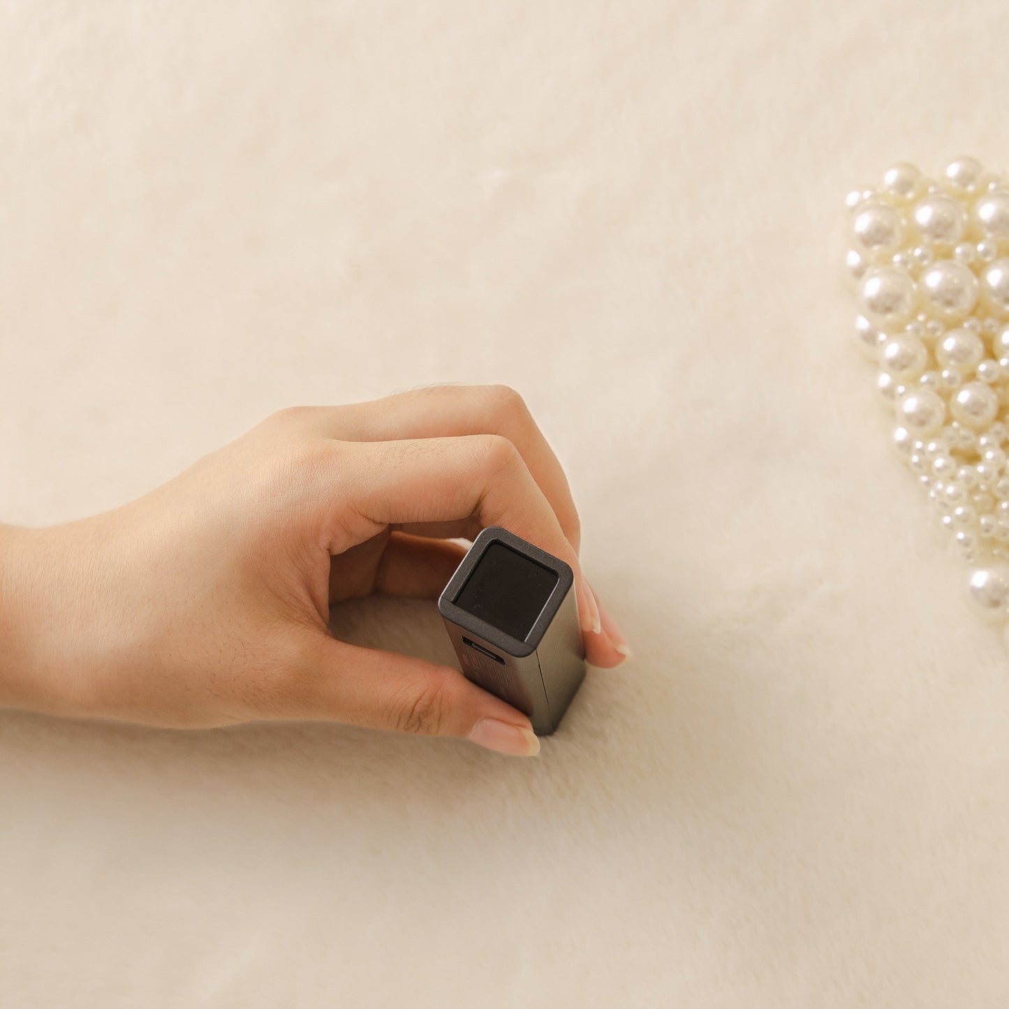 Hand holding a rectangular black object on a soft beige fabric surface, with pearls in the background.