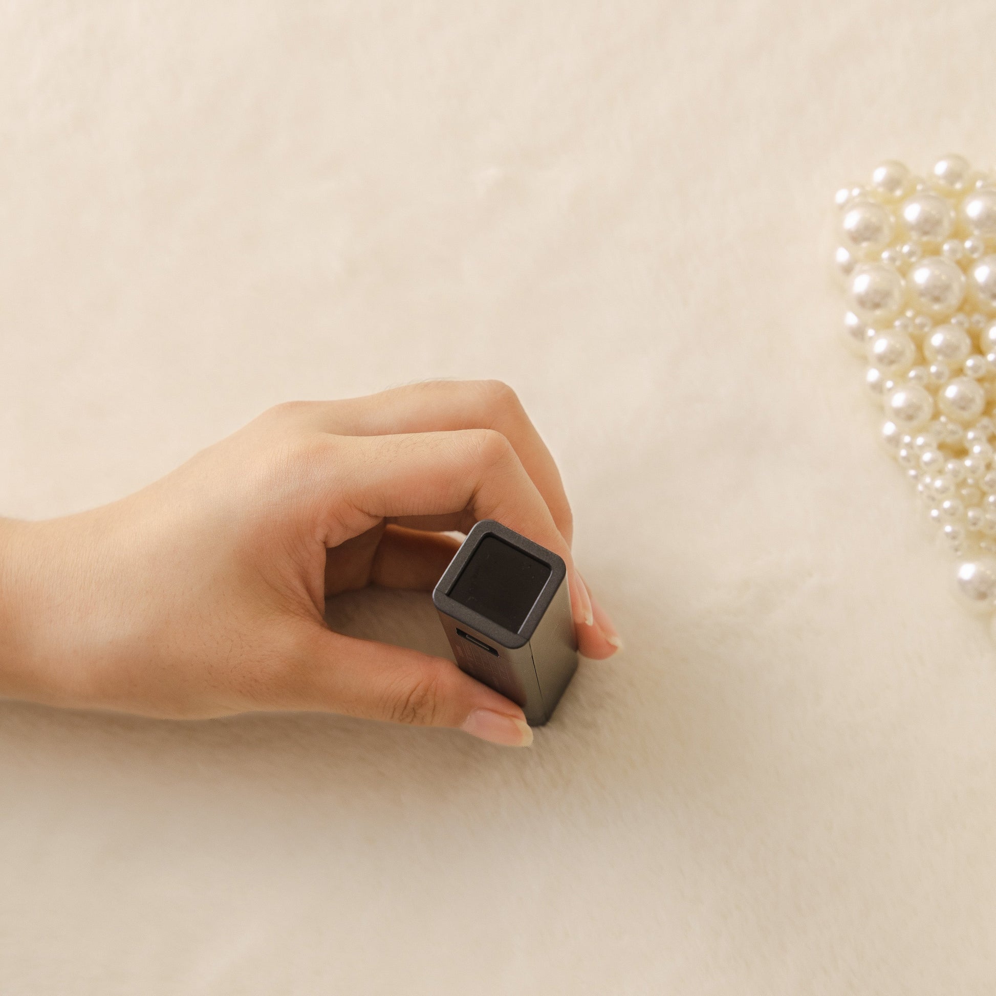 Hand holding a rectangular black object on a soft beige fabric surface, with pearls in the background.