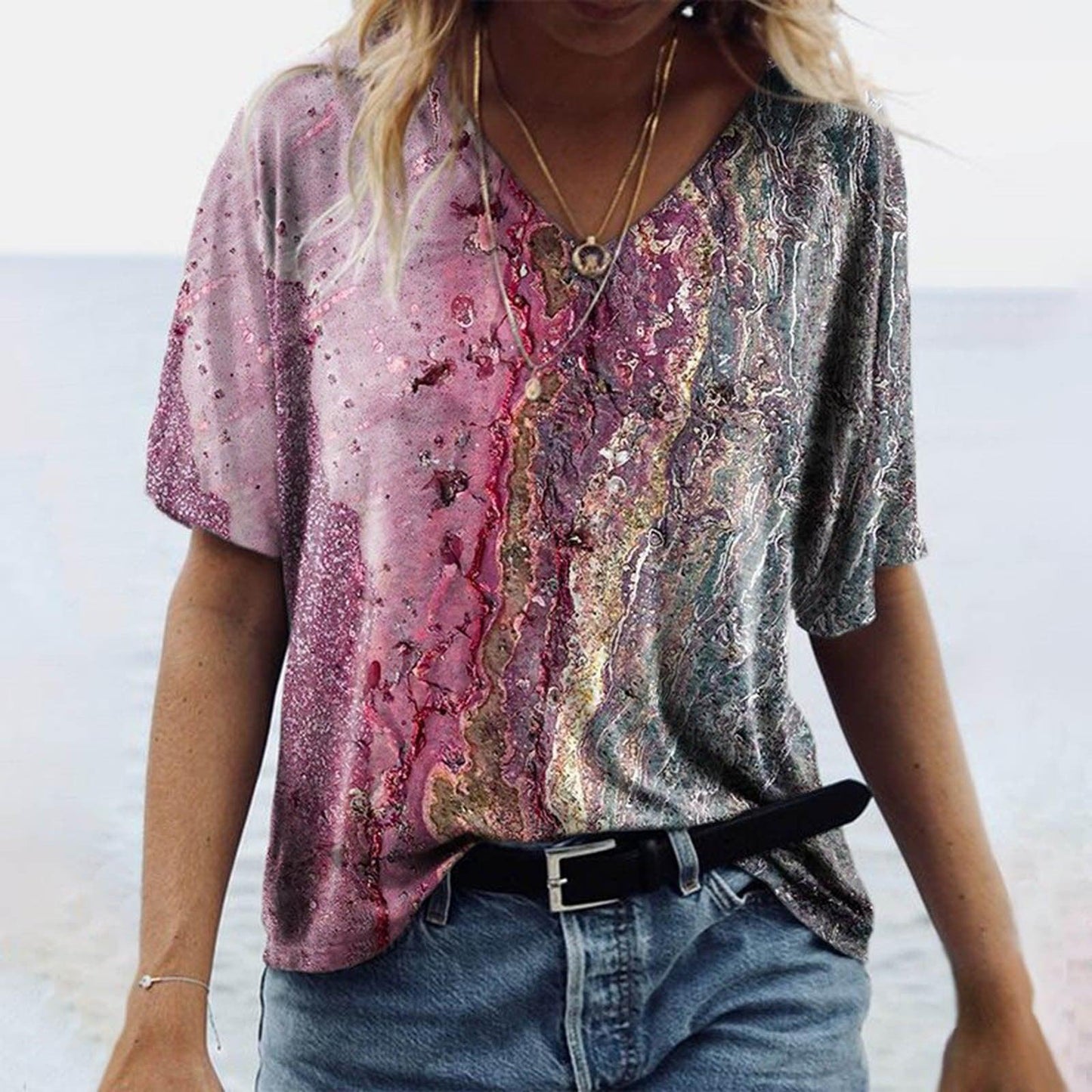 Woman wearing Women's Spring Glitzy V-Neck Short Sleeved Marble Print Top with multicolor tie-dye effect, standing on beach.