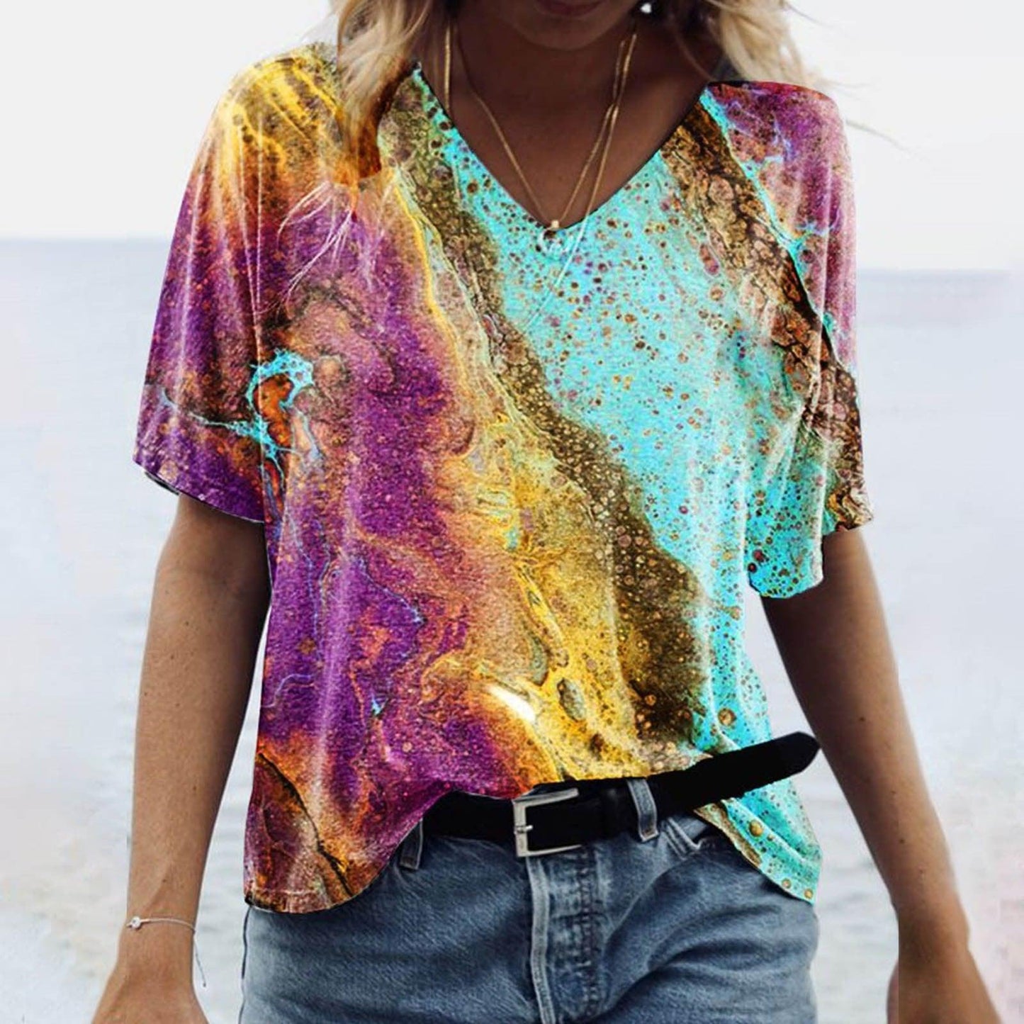 Woman wearing a Women's Spring Glitzy V-Neck Short Sleeved Marble Print Top in colorful tie-dye marble pattern with a black belt, beach background.
