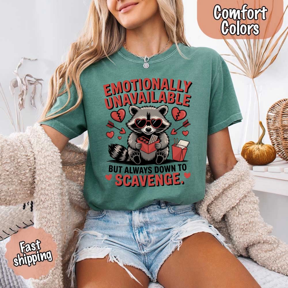 Woman wearing Comfort Colors Emotionally Unavailable Raccoon T-Shirts, featuring a moody raccoon print with funny text, cozy home setting.