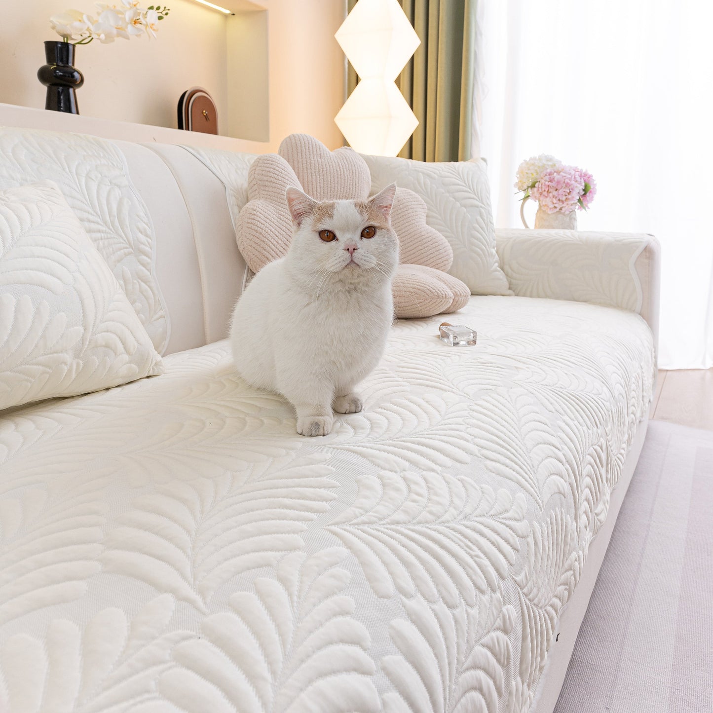 White cat sitting on a sofa with Sofa Cover for Leather & Fabric Couches, 3D Leaf Embossed Quilted Furniture Covers in a living room.