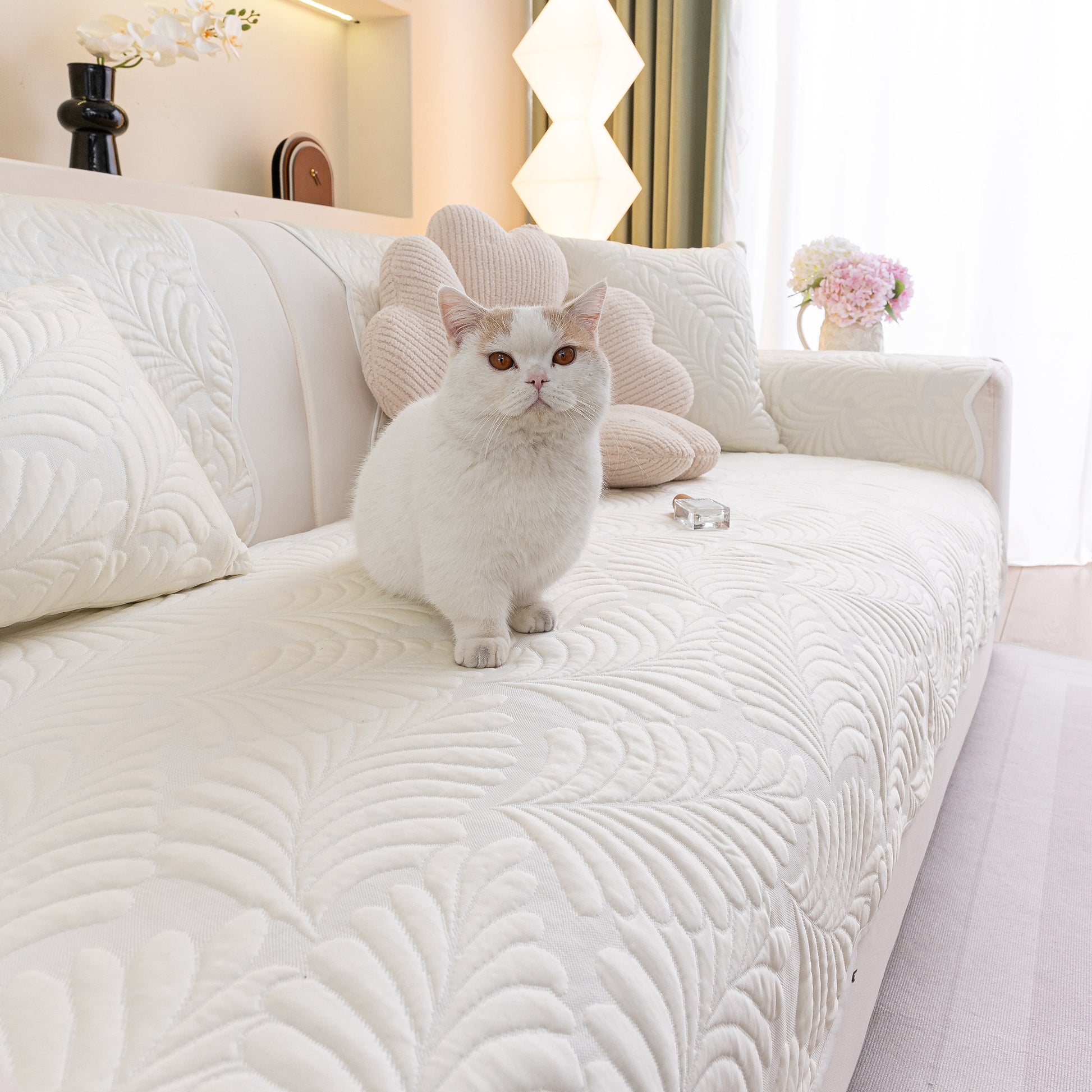White cat sitting on a sofa with Sofa Cover for Leather & Fabric Couches, 3D Leaf Embossed Quilted Furniture Covers in a living room.