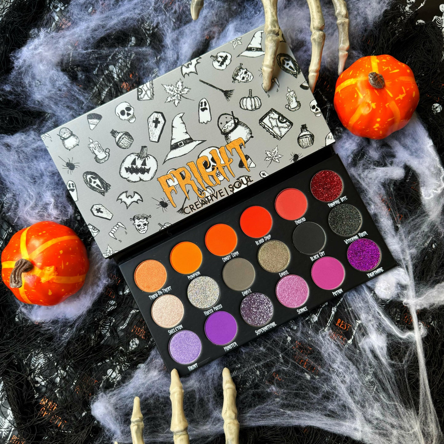 FRIGHT Eyeshadow Palette surrounded by Halloween pumpkins and cobwebs, showcasing vibrant purples, reds, oranges, and grays.