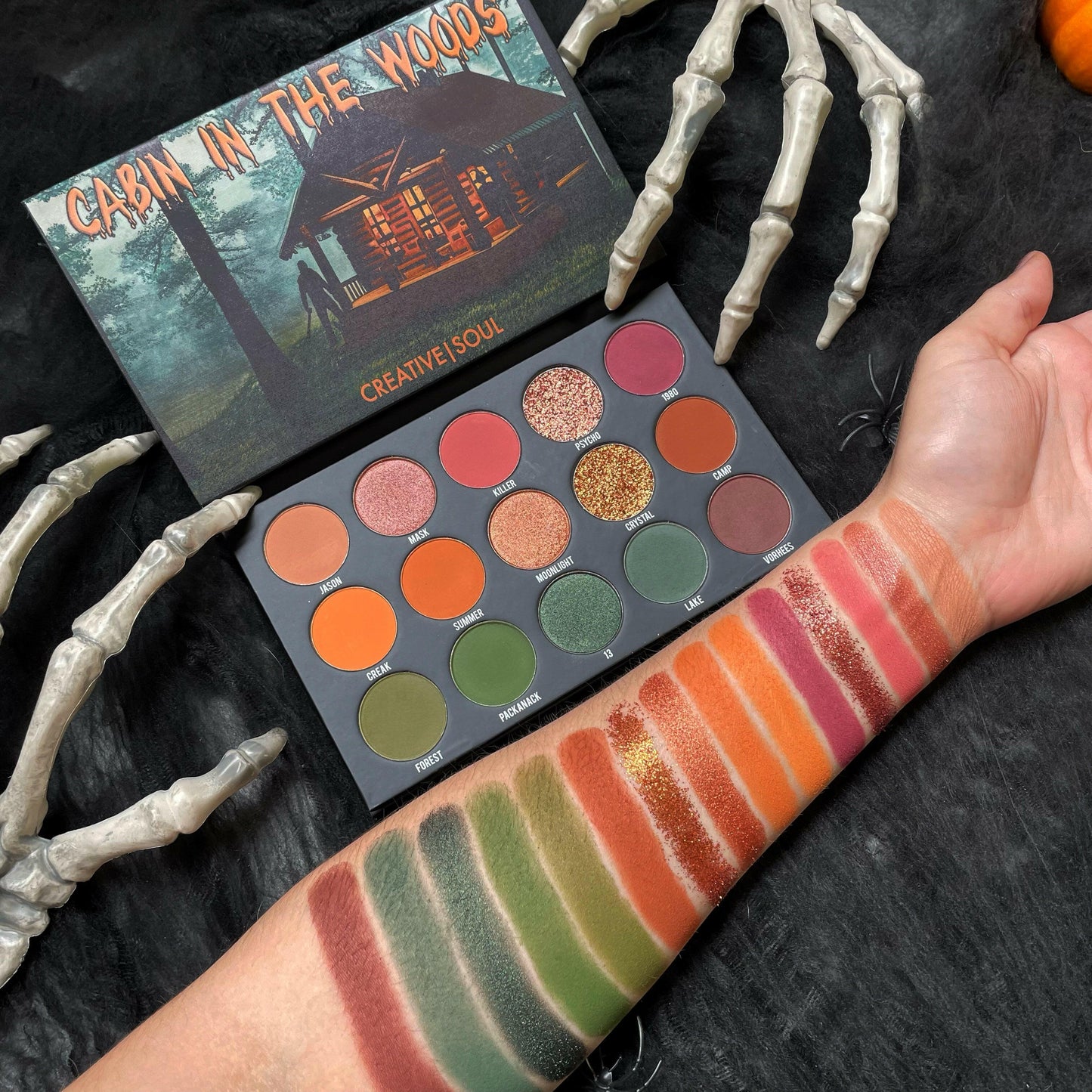 Cabin In The Woods Eyeshadow Palette with swatches of mauve, pink, orange, and green shades on an arm next to skeleton hands.