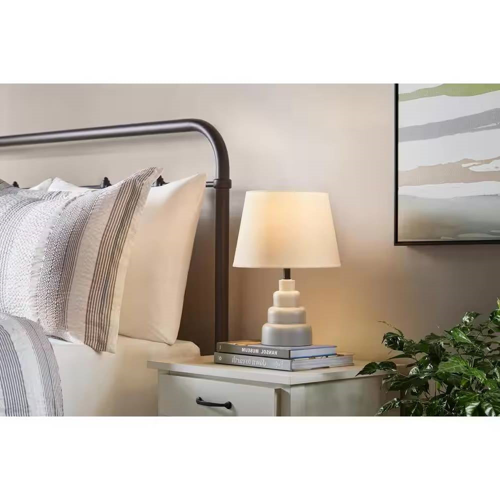 Small Grey Desk Light Table Lamp with White Fabric Shade for Bedside Nightstand on a bedroom nightstand next to a bed.