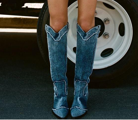 Woman wearing Handcrafted Distressed Denim Knee-High Boots standing in front of a vehicle tire on pavement.
