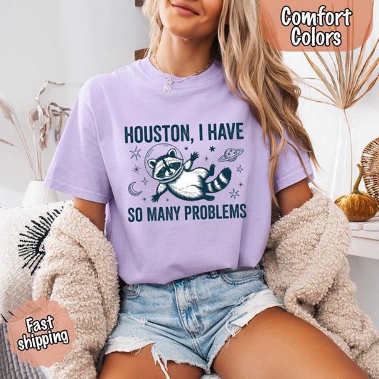 Houston I Have So Many Problems Raccoon Comfort Colors T-Shirts featuring a space raccoon design, worn by a woman sitting comfortably.