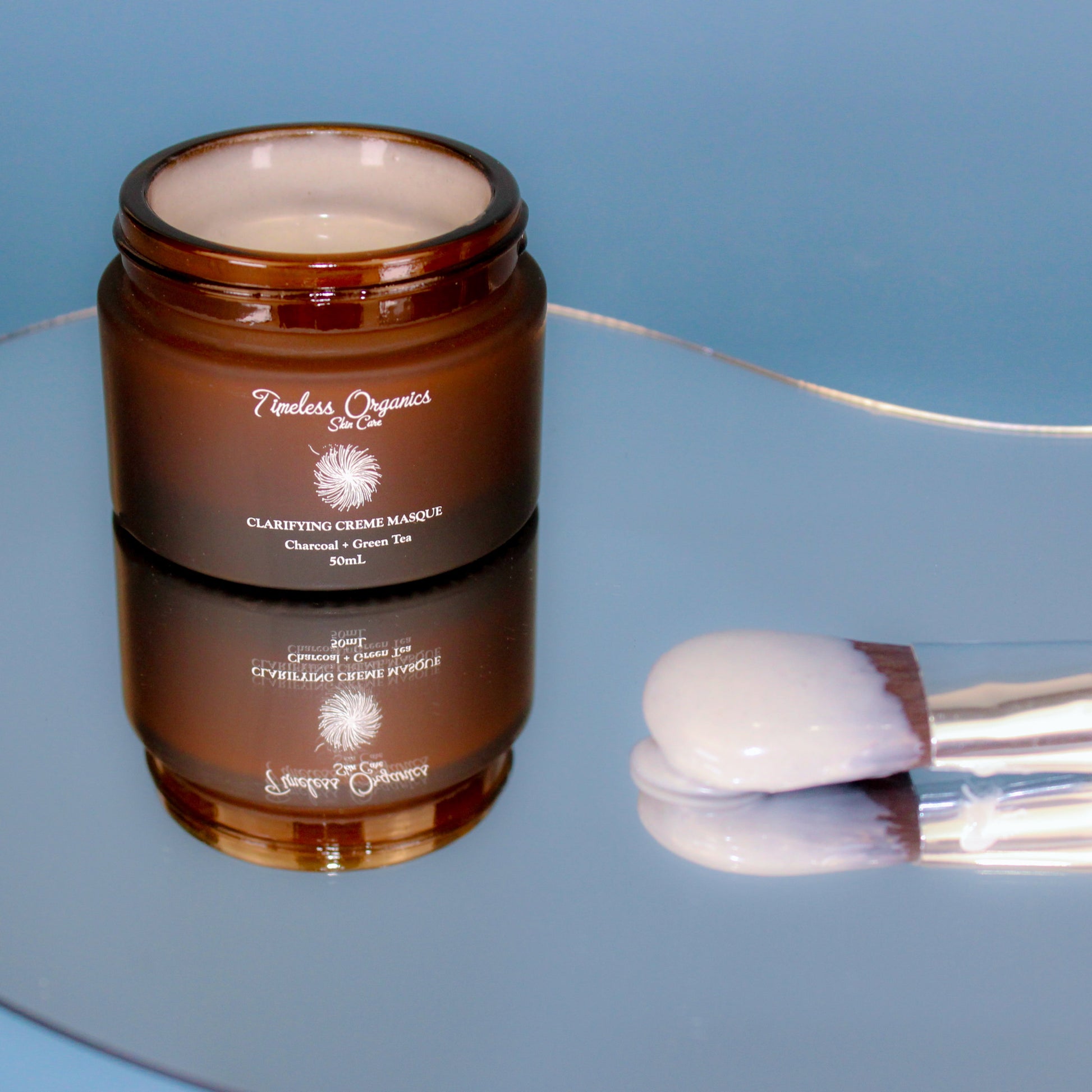 Clarifying Charcoal Creme Masque with applicator brush on reflective surface, 50mL jar