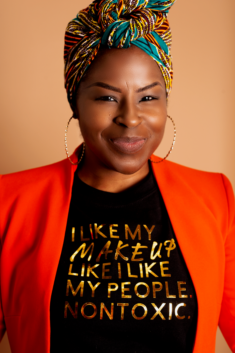Person wearing black 'I Like My Makeup Like...' Tee with gold text, orange jacket, and patterned headscarf.
