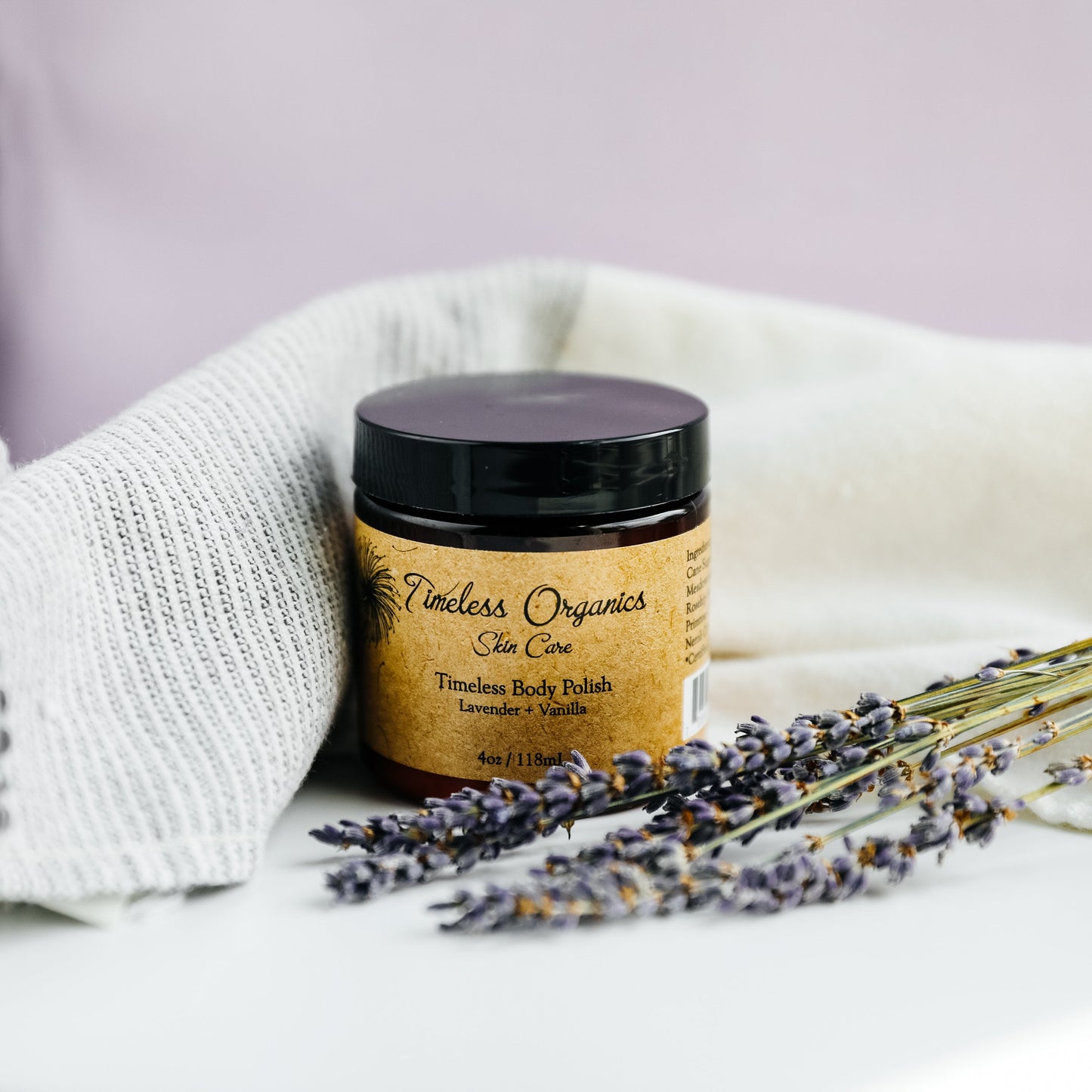 Body Polish - Lavender + Vanilla in jar with lavender sprigs, offering gentle exfoliation for soft skin.