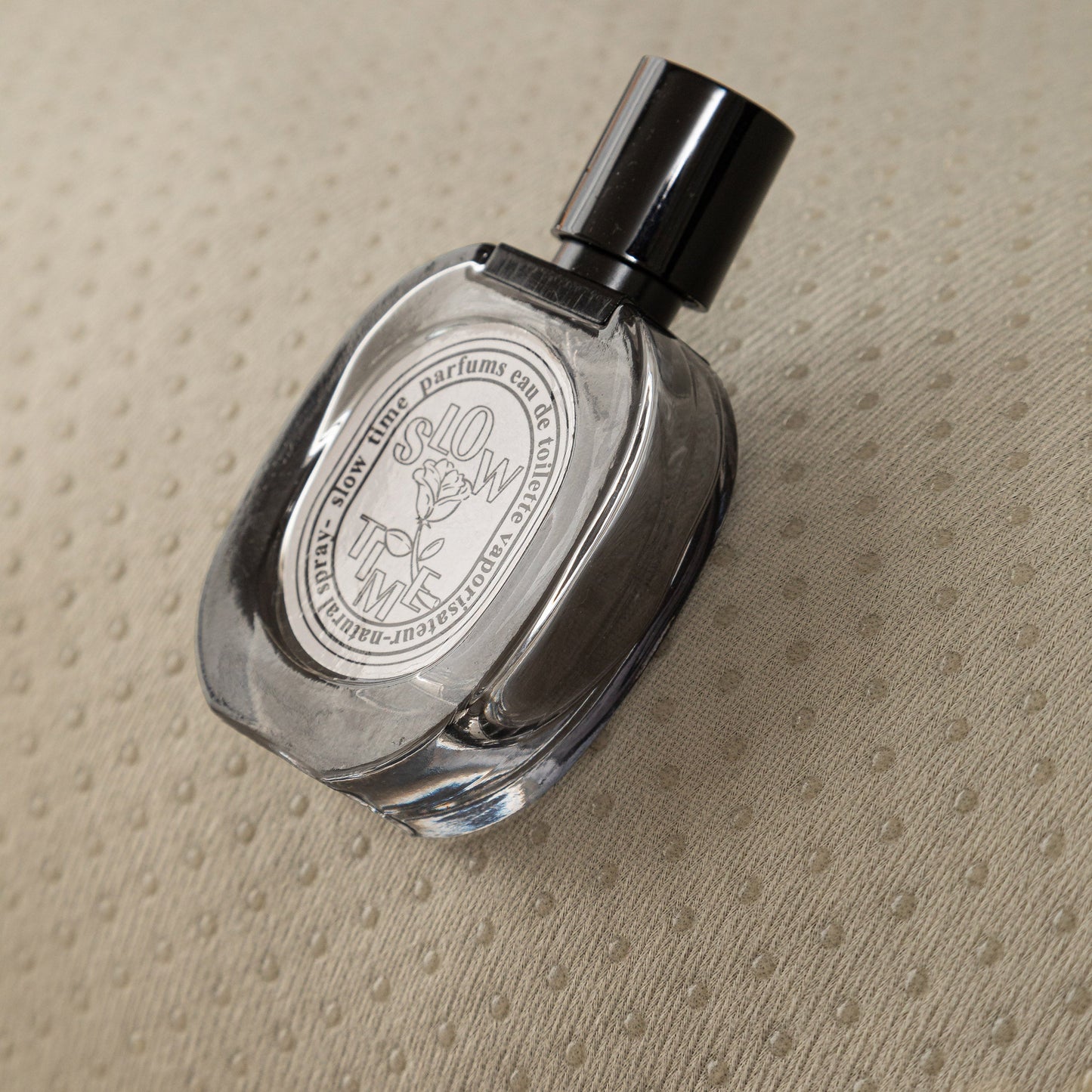Transparent glass perfume bottle with black cap on a textured beige surface.