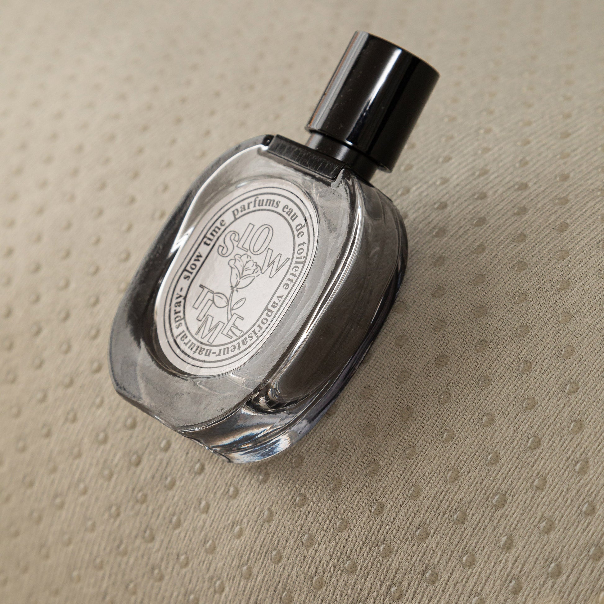 Transparent glass perfume bottle with black cap on a textured beige surface.