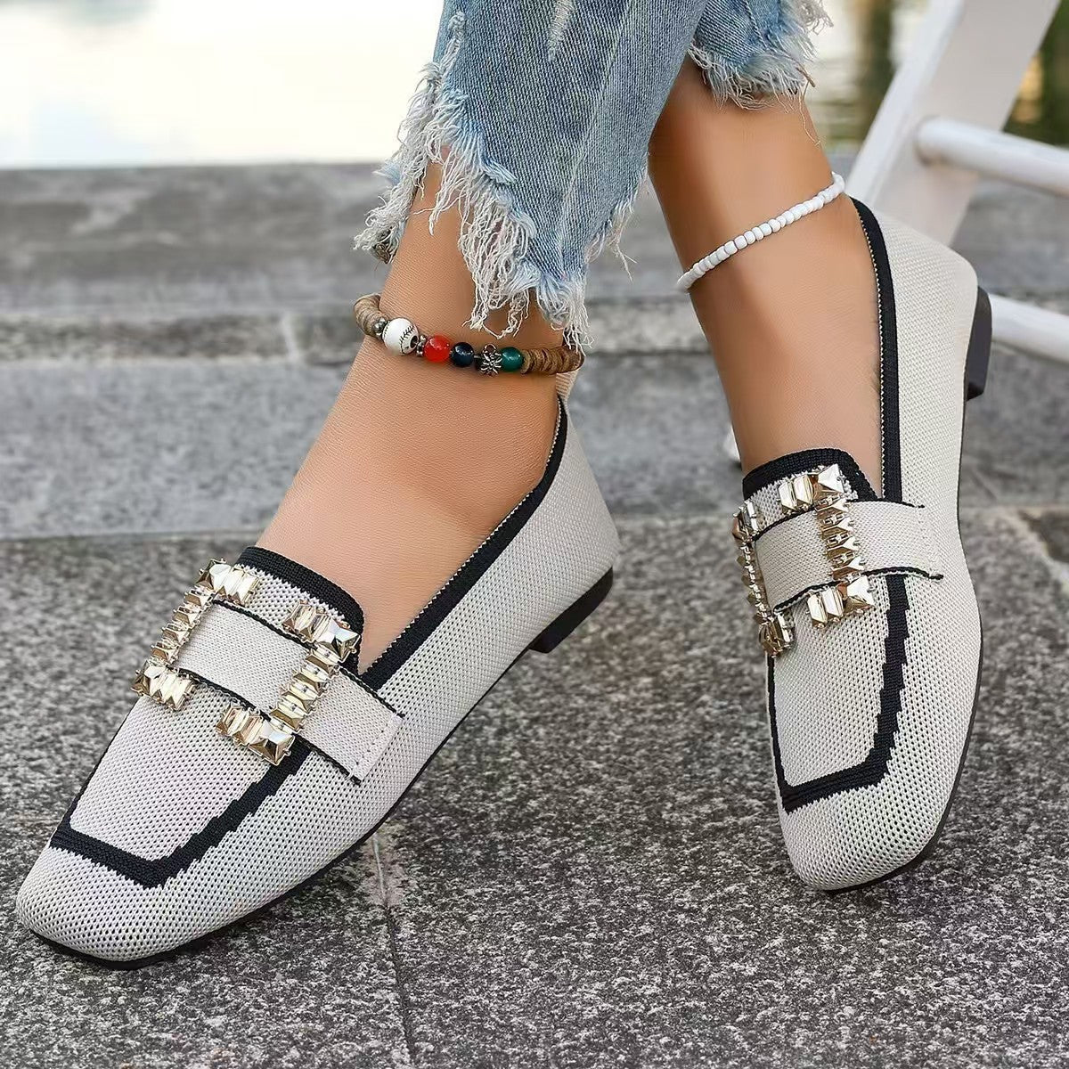 Close-up of stylish white loafers with jeweled buckles, worn with distressed jeans and ankle bracelet on pavement.