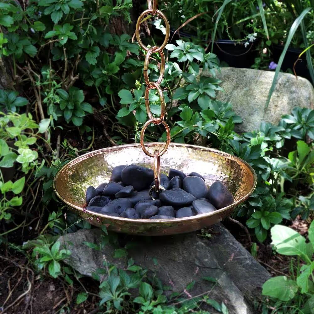 Hand Hammered Copper Polished Rain Chain Basin - 16.5 inch Diameter with black stones in a lush garden setting