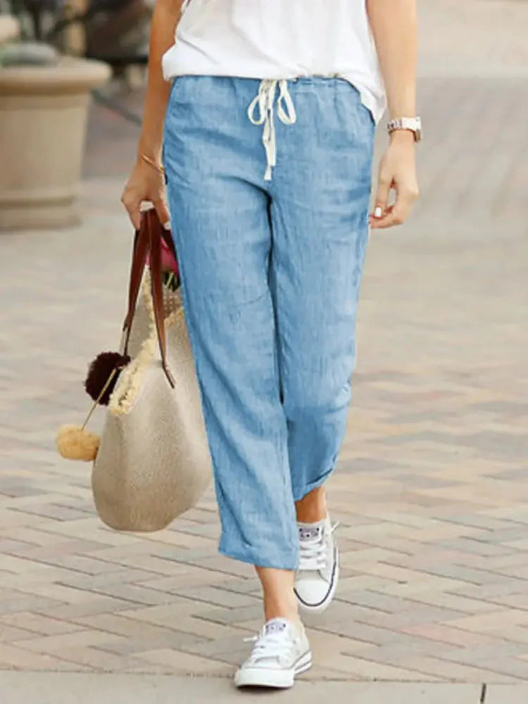 Person walking wearing light blue drawstring casual pants and white sneakers, holding a straw bag with pom-poms.