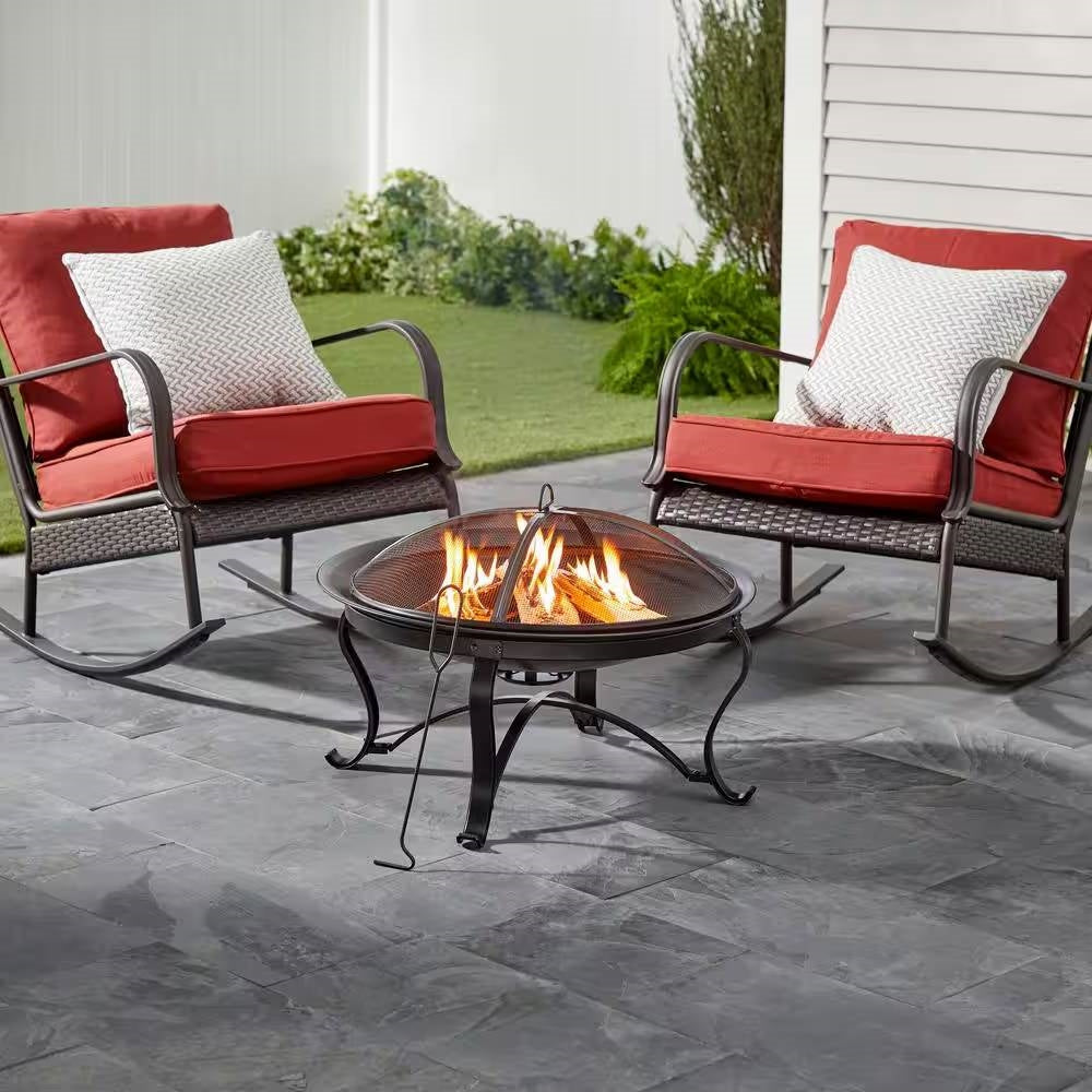 30-inch Round Steel Outdoor Fire Pit with Mesh Cover and Poker in Bronze set on patio between two red-cushioned chairs.