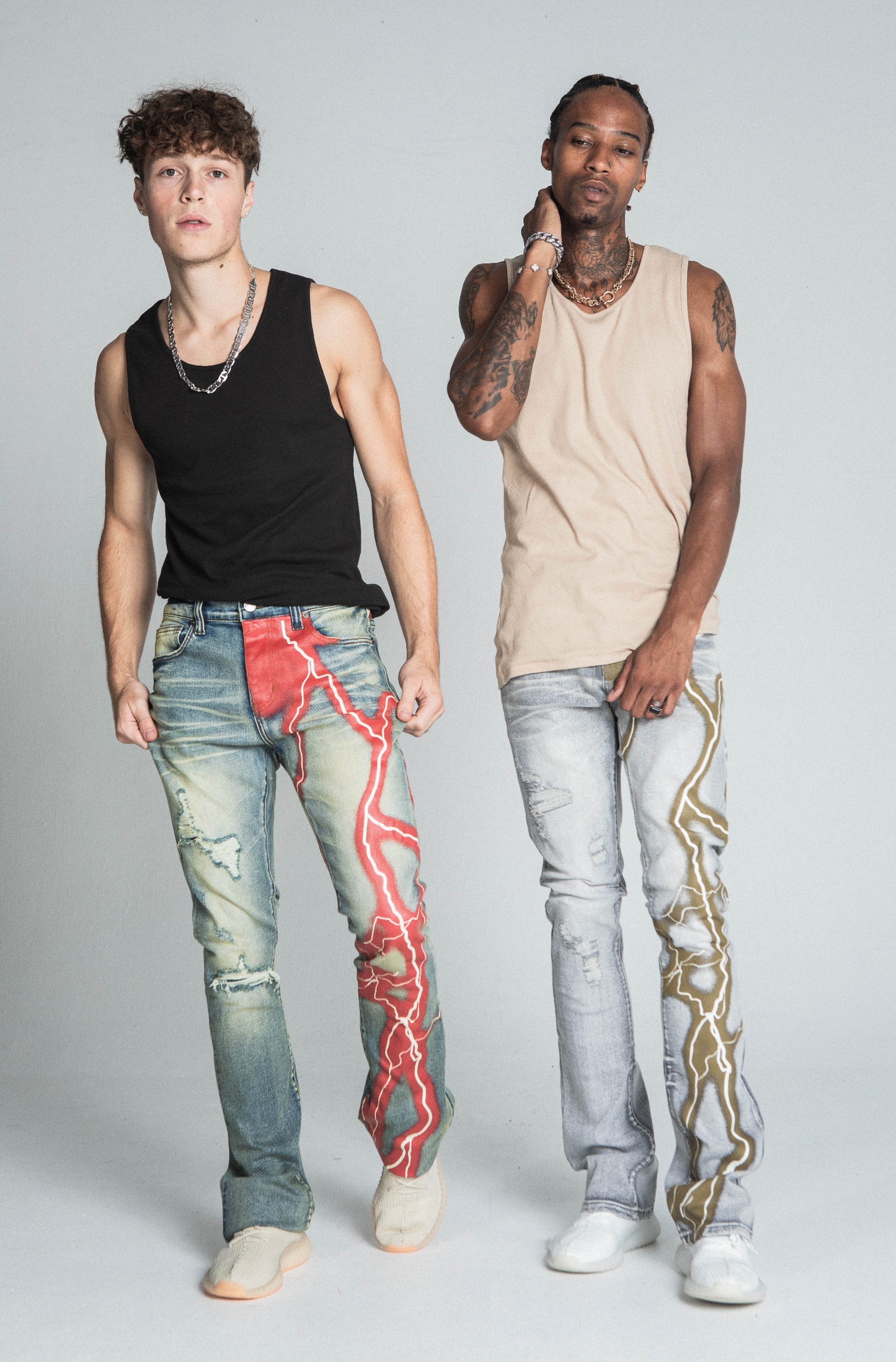 Two models wearing AJ24SK LIGHTNING STACKED JEANS with colorful lightning design in a studio setting.