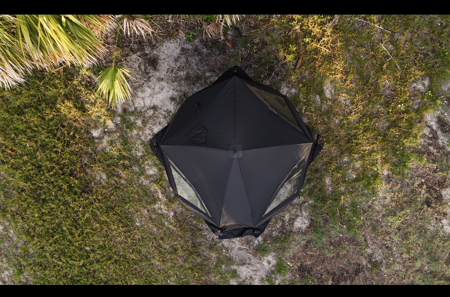 Aerial view of Nova 6 Sauna Tent (6-person; tent only) set up on grassy terrain, showcasing its hexagonal shape and black color.