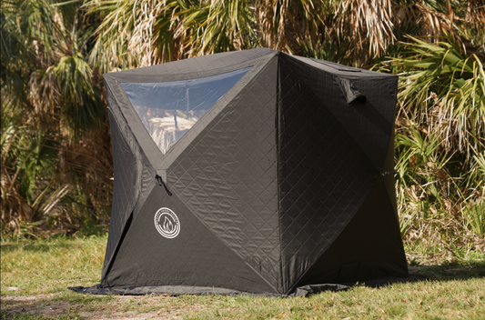 Black Mini Cube Sauna Tent (2-person; tent only) set up outdoors in grassy area with palm trees in the background.