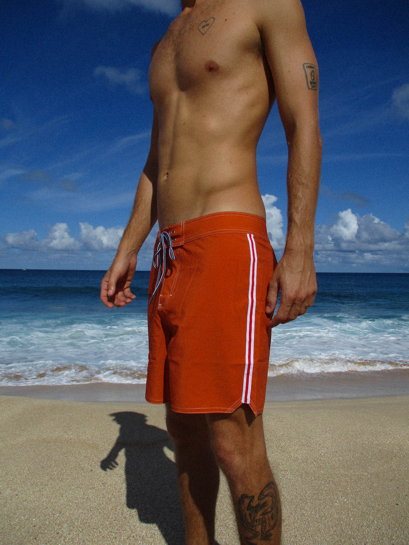 Model wearing the classic boardshort - sunset in burnt orange by the ocean.