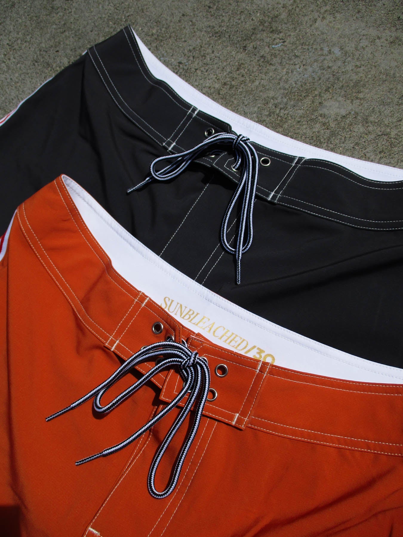 Two pairs of boardshorts, one in burnt orange and one in black, showcasing the classic boardshort - sunset with drawstring eyelets.