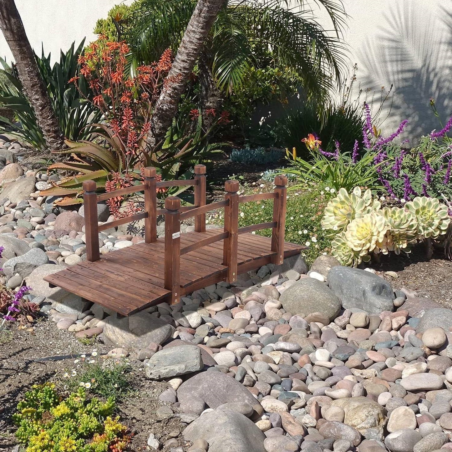 Outdoor 5-foot Solid Fir Wood Garden Bridge in Rustic Brown Finish over decorative garden rocks and plants.
