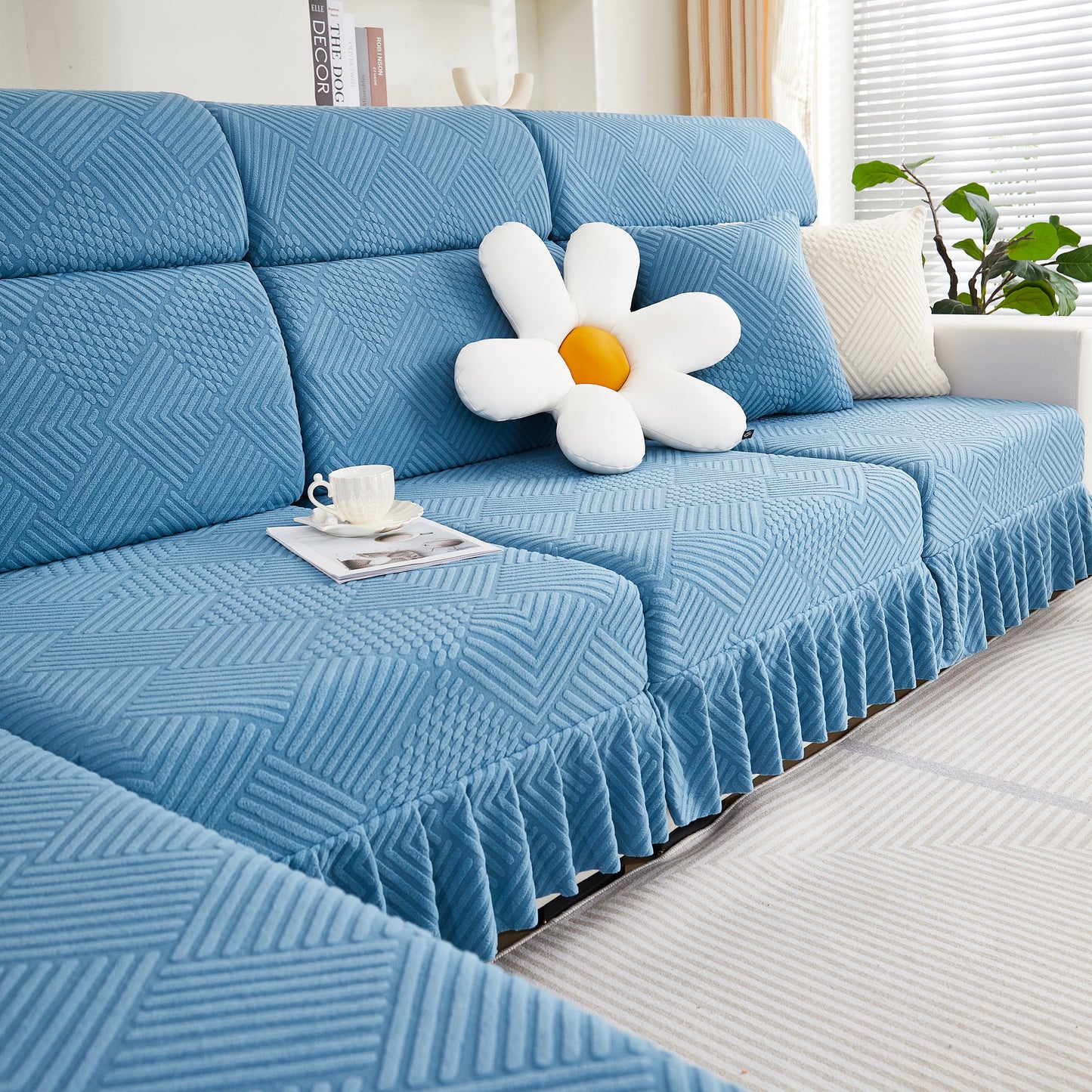 Blue stretch sofa covers with a skirt on a 4-seater couch, featuring a daisy pillow and coffee cup, for a modern living room look.