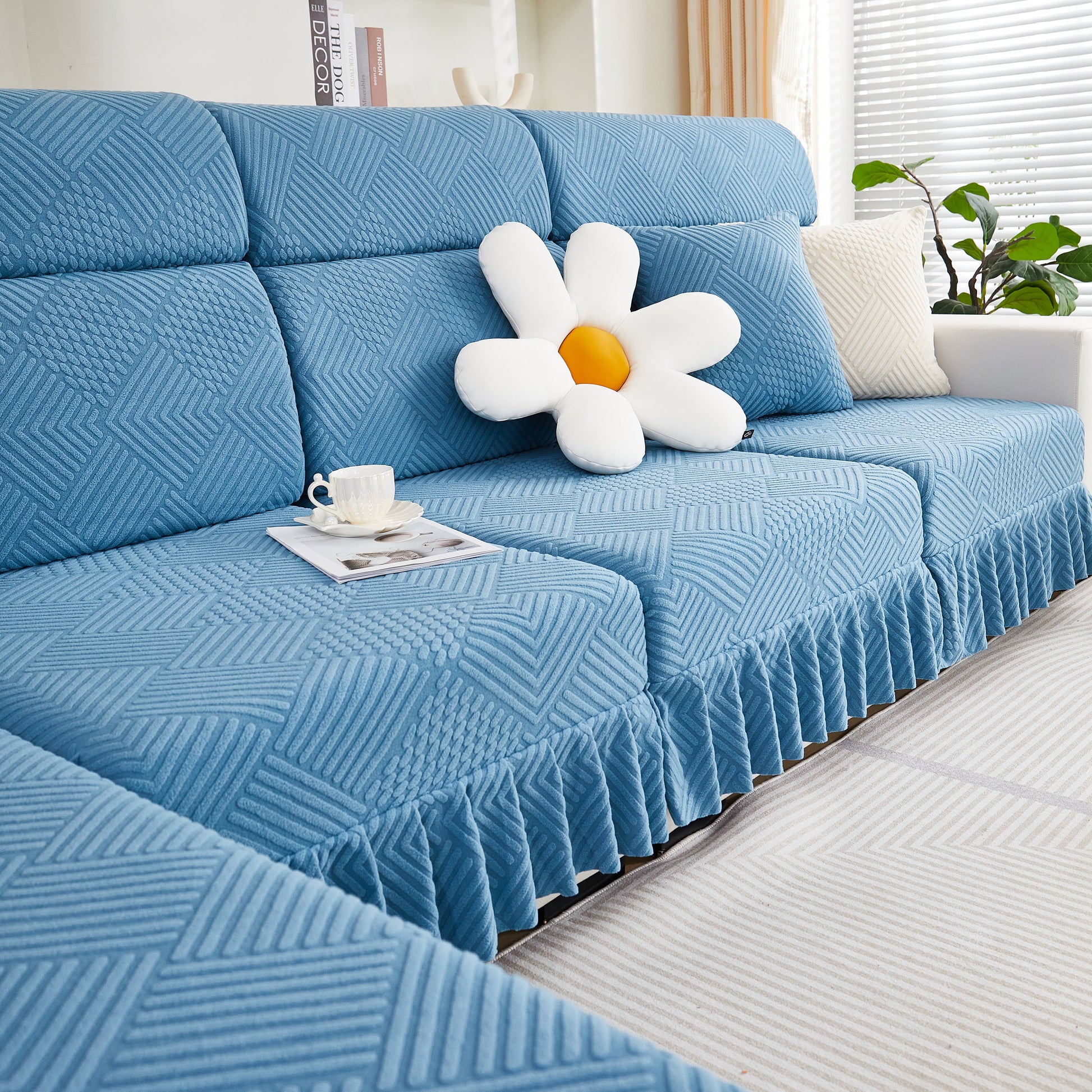 Blue stretch sofa covers with a skirt on a 4-seater couch, featuring a daisy pillow and coffee cup, for a modern living room look.