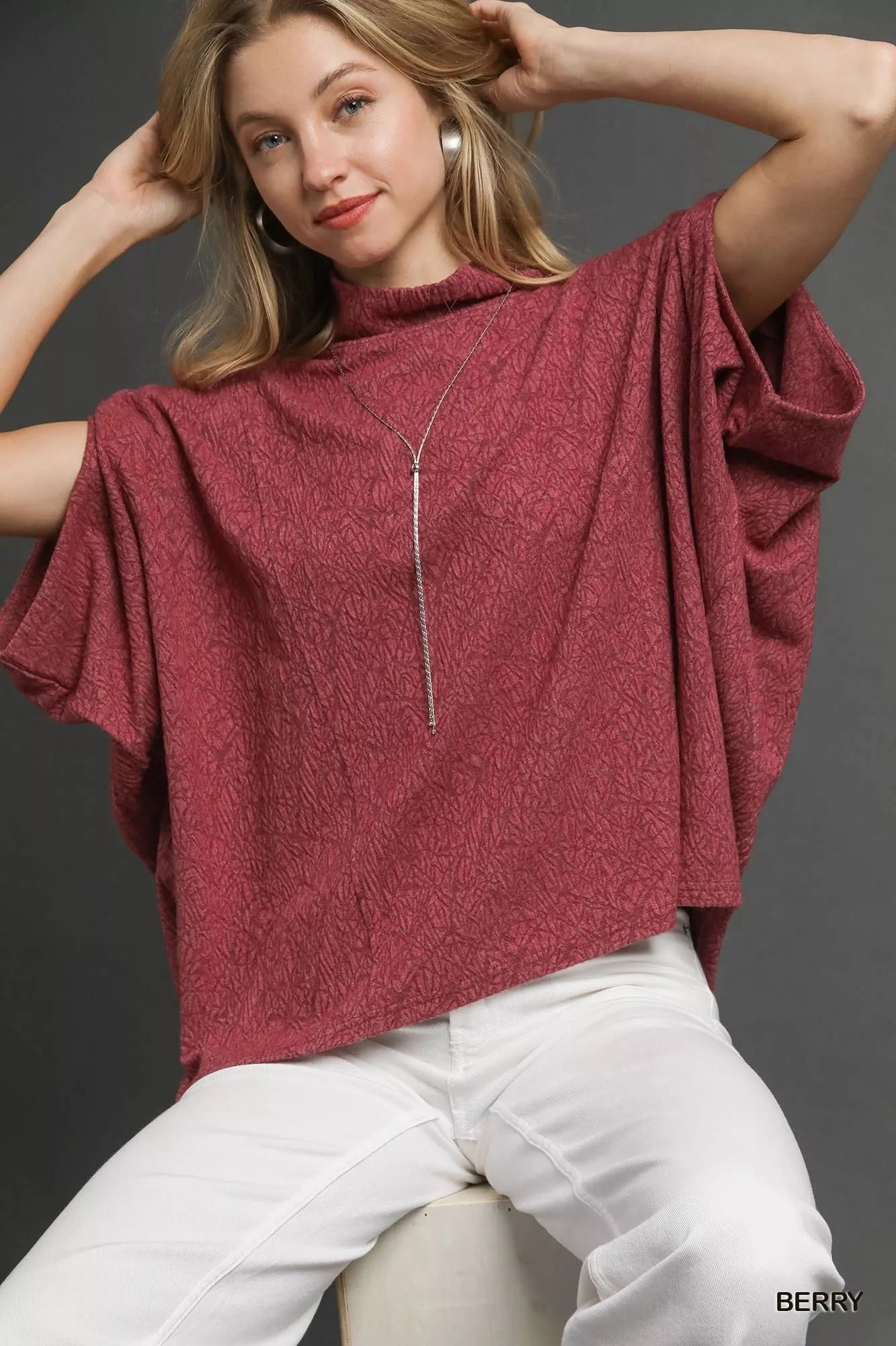 Woman wearing Umgee Textured Mock Neck Relaxed Top in berry color with dolman sleeves and side slits, styled with white pants.