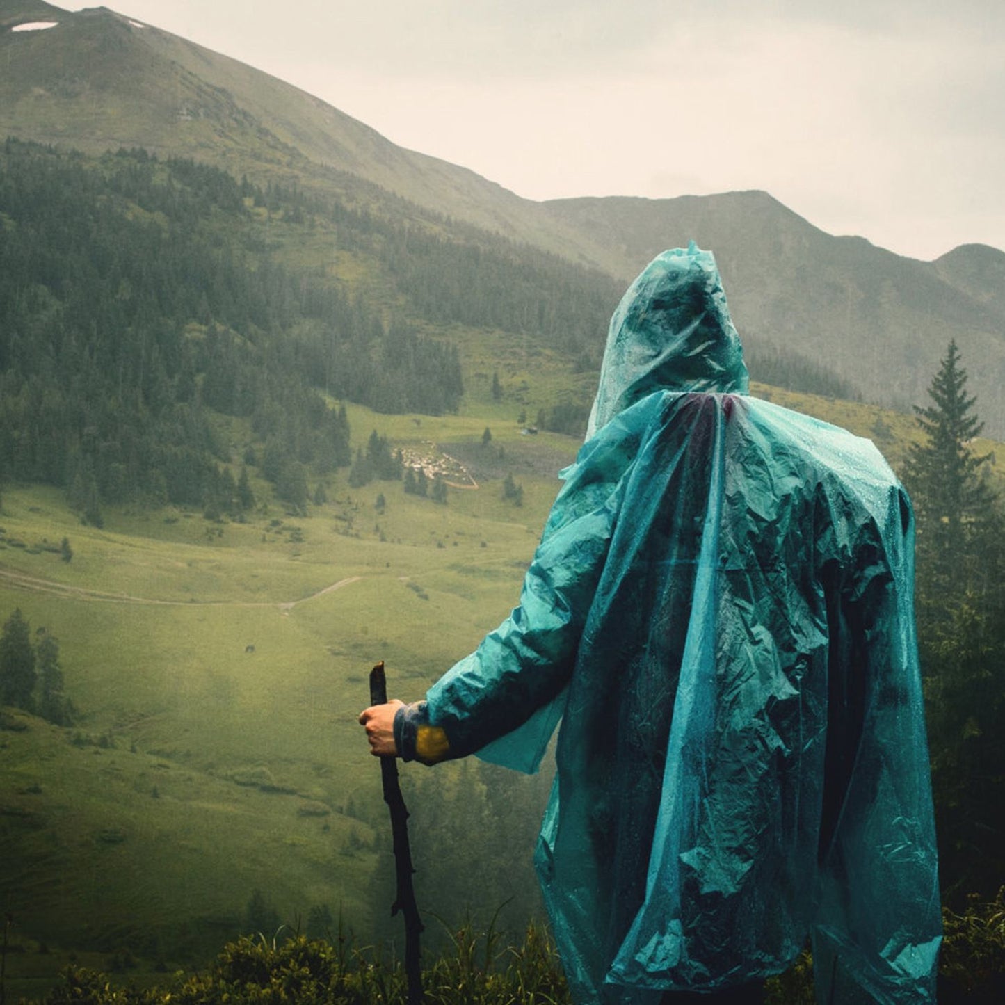 Hiking in the rain with a blue poncho in scenic mountain landscape, showcasing essentials from a 125Pcs Survival Kit for outdoor adventures.