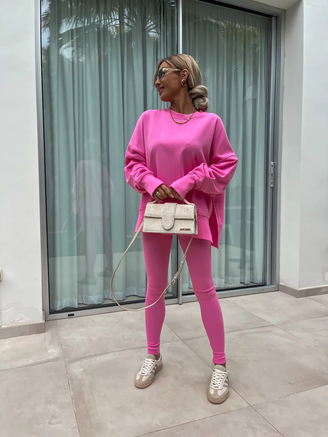 Woman wearing Women’s Casual Sweater Suit – Loose Split Top & Fitted Trousers Set in pink, standing outside by a window holding a small bag.