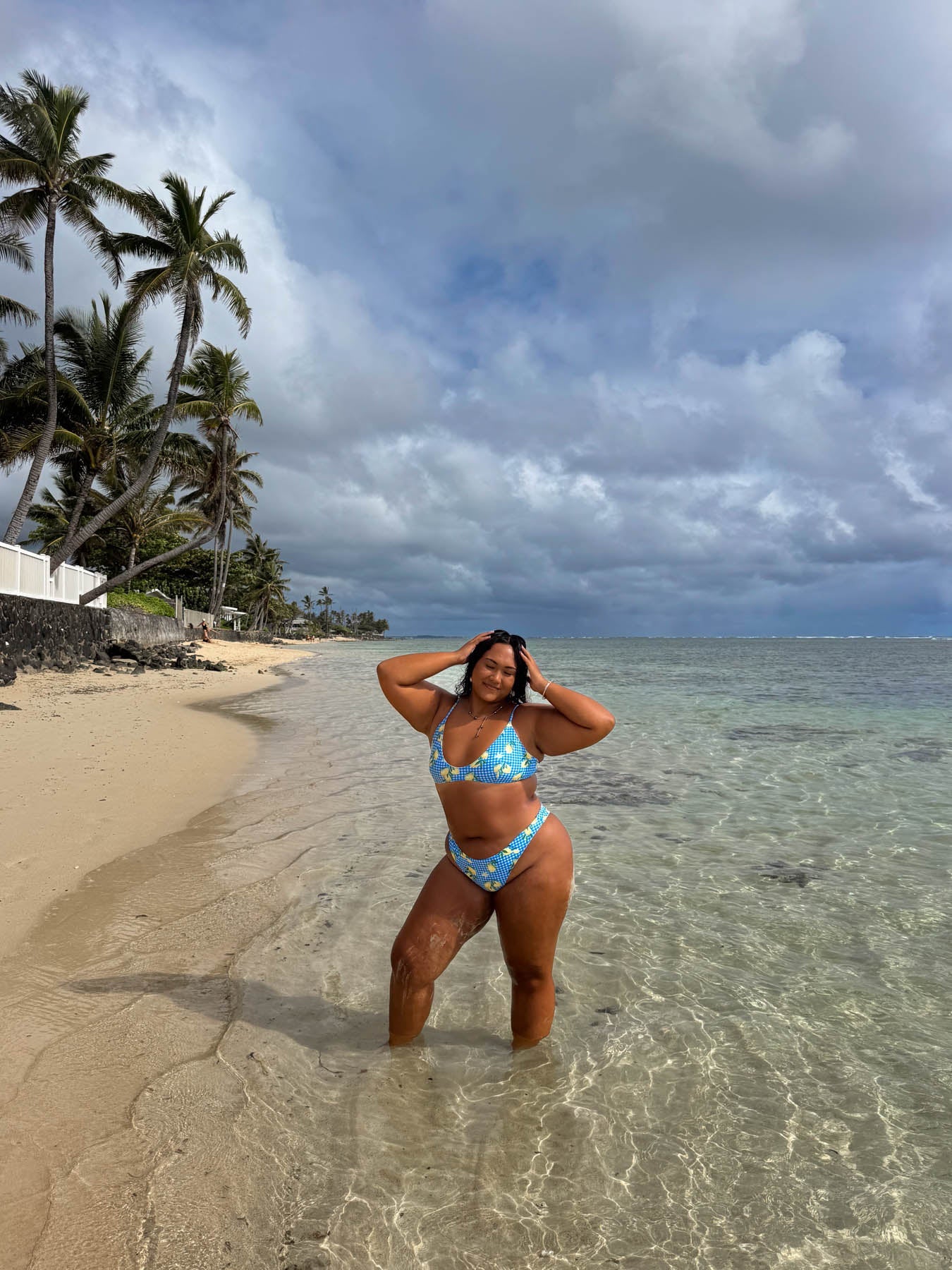Woman wearing sky bottom - amalfi bikini standing in clear ocean water with cloudy sky and palm trees in the background.