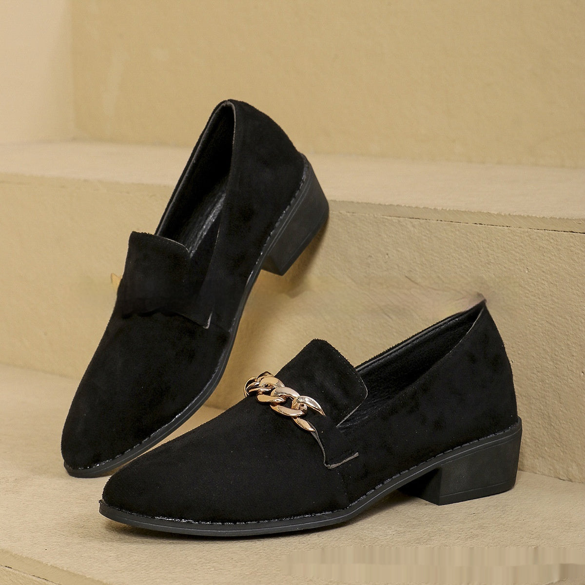 Pair of black suede loafers with chain detail on a beige background