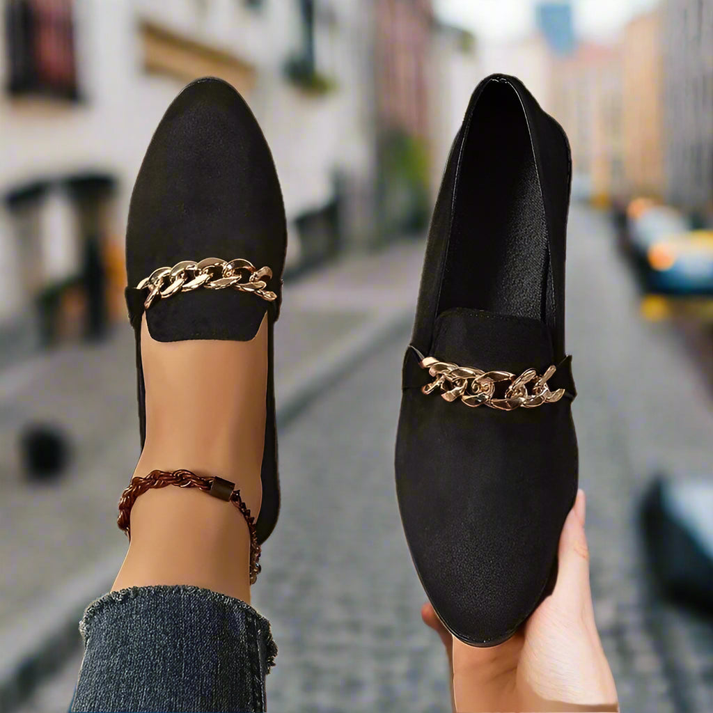 Stylish black flat shoes with gold chain detail on cobblestone street.