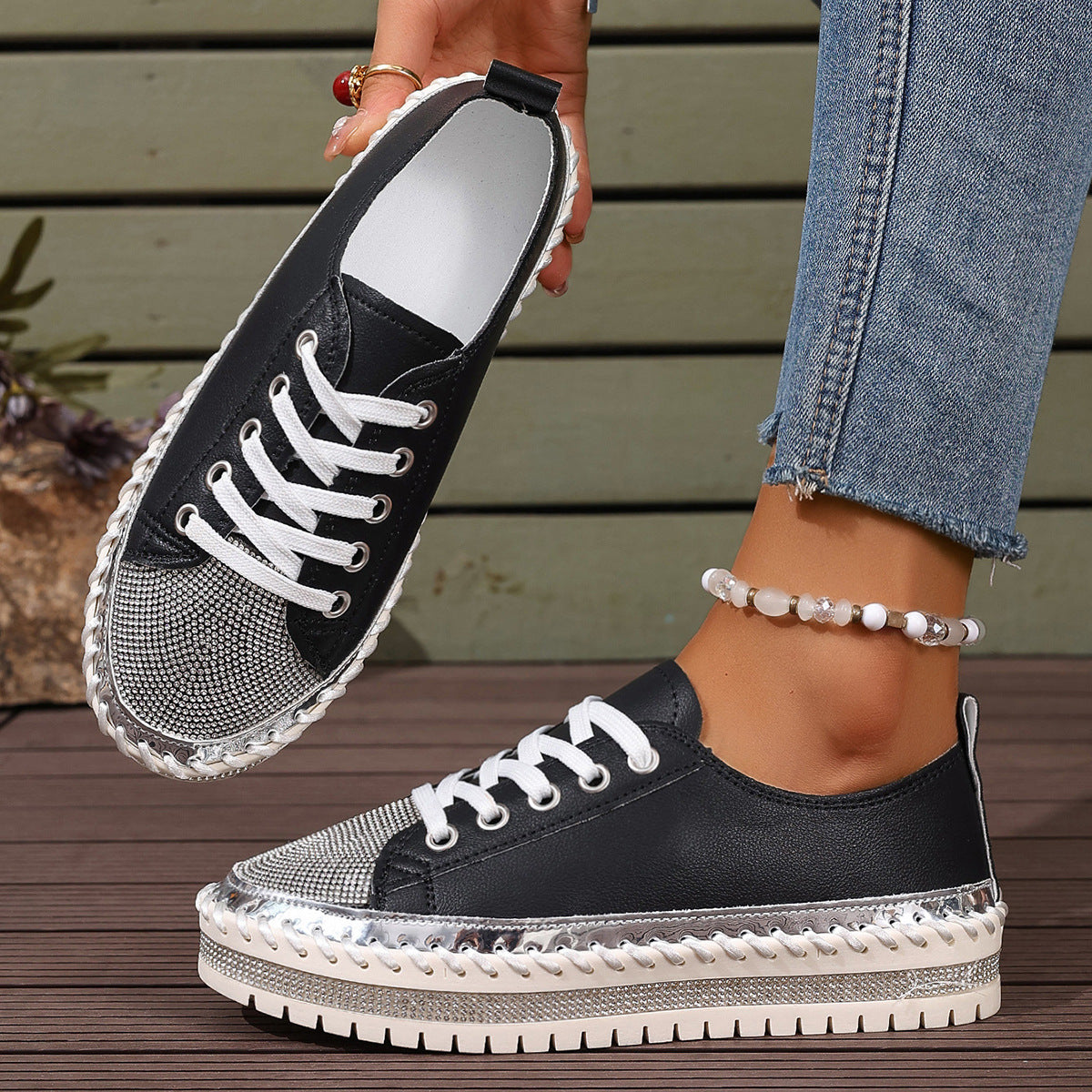 Black sneakers with metallic accents and chunky sole, worn with jeans and an anklet on wooden deck background.