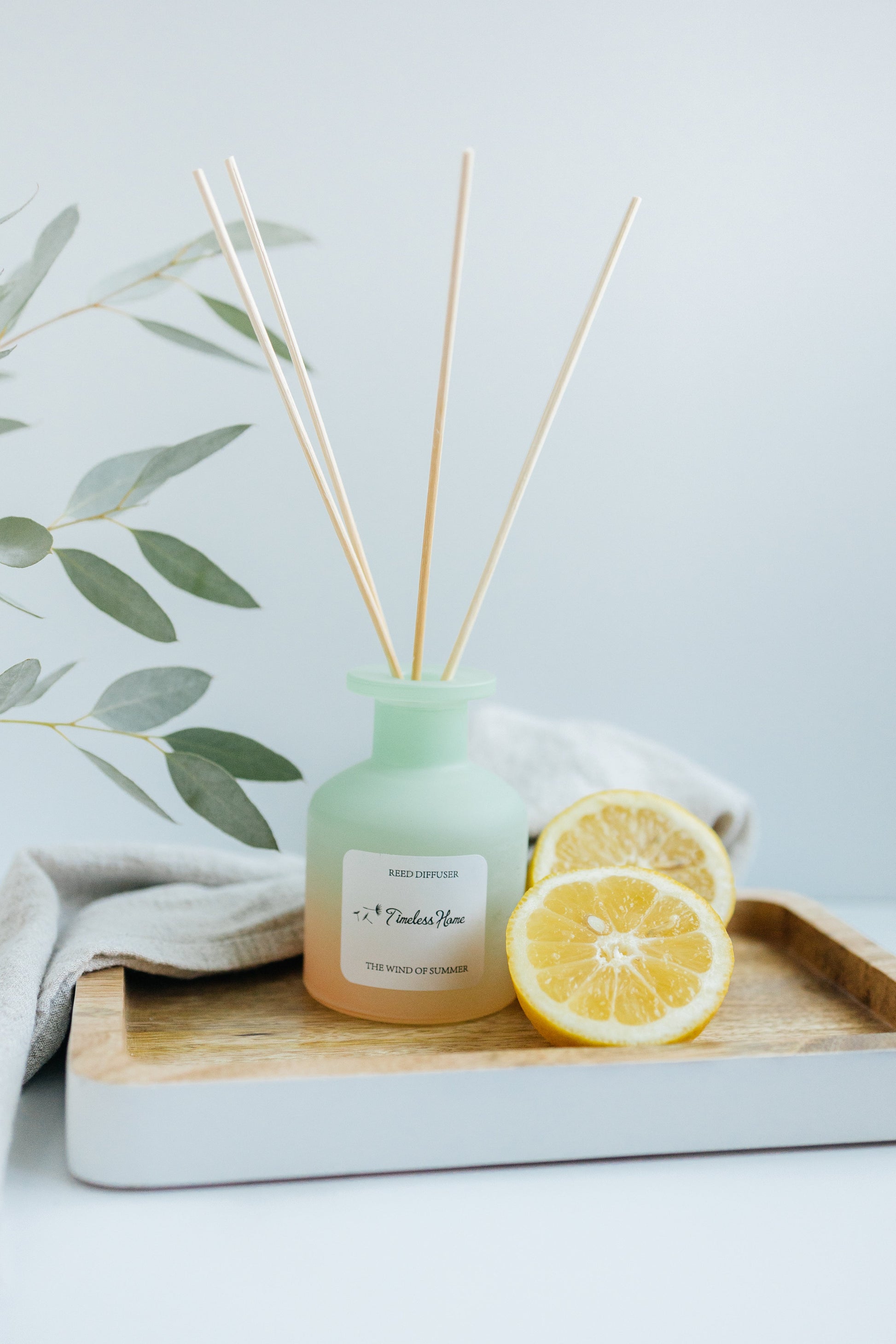 Reed Diffuser - The Wind of Summer with natural reed sticks and frosted glass vessel, surrounded by fresh lemons on a wooden tray.