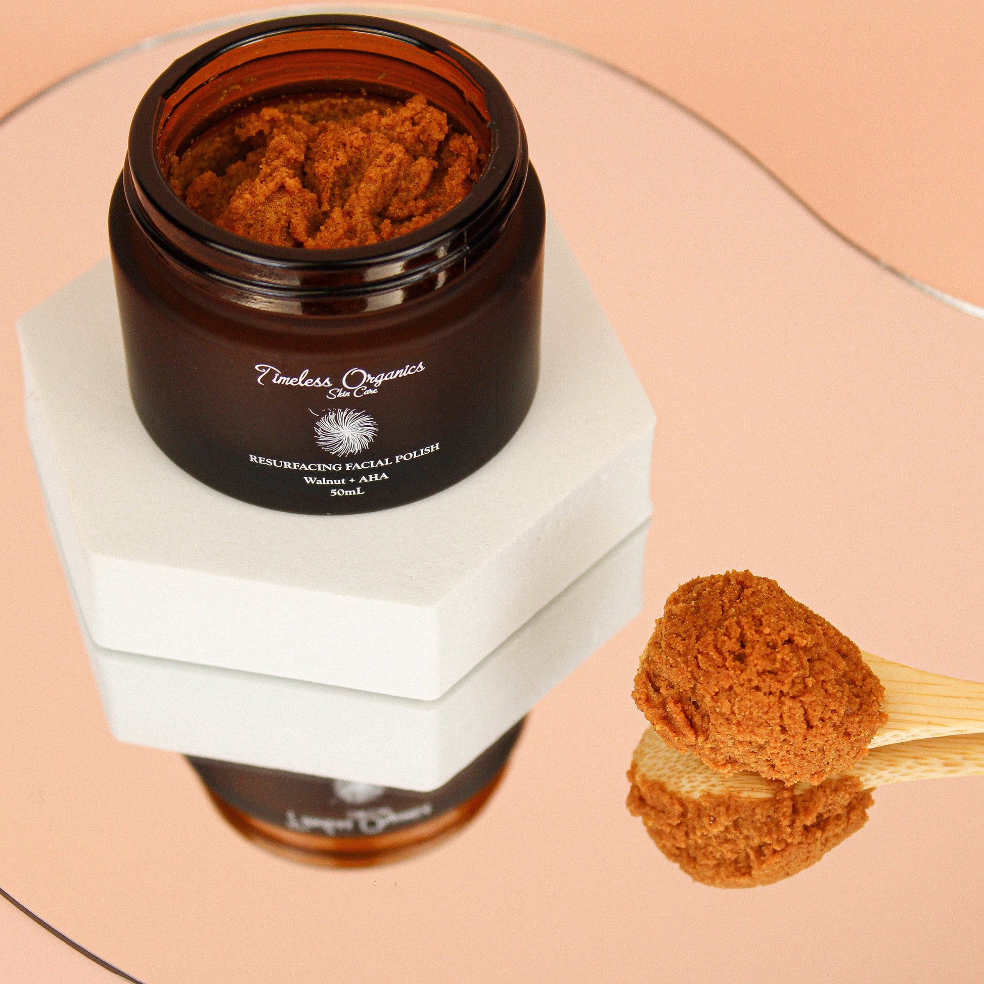 Open jar of Resurfacing Facial Polish - Walnut + AHA on a mirrored surface with a small scoop on the side.