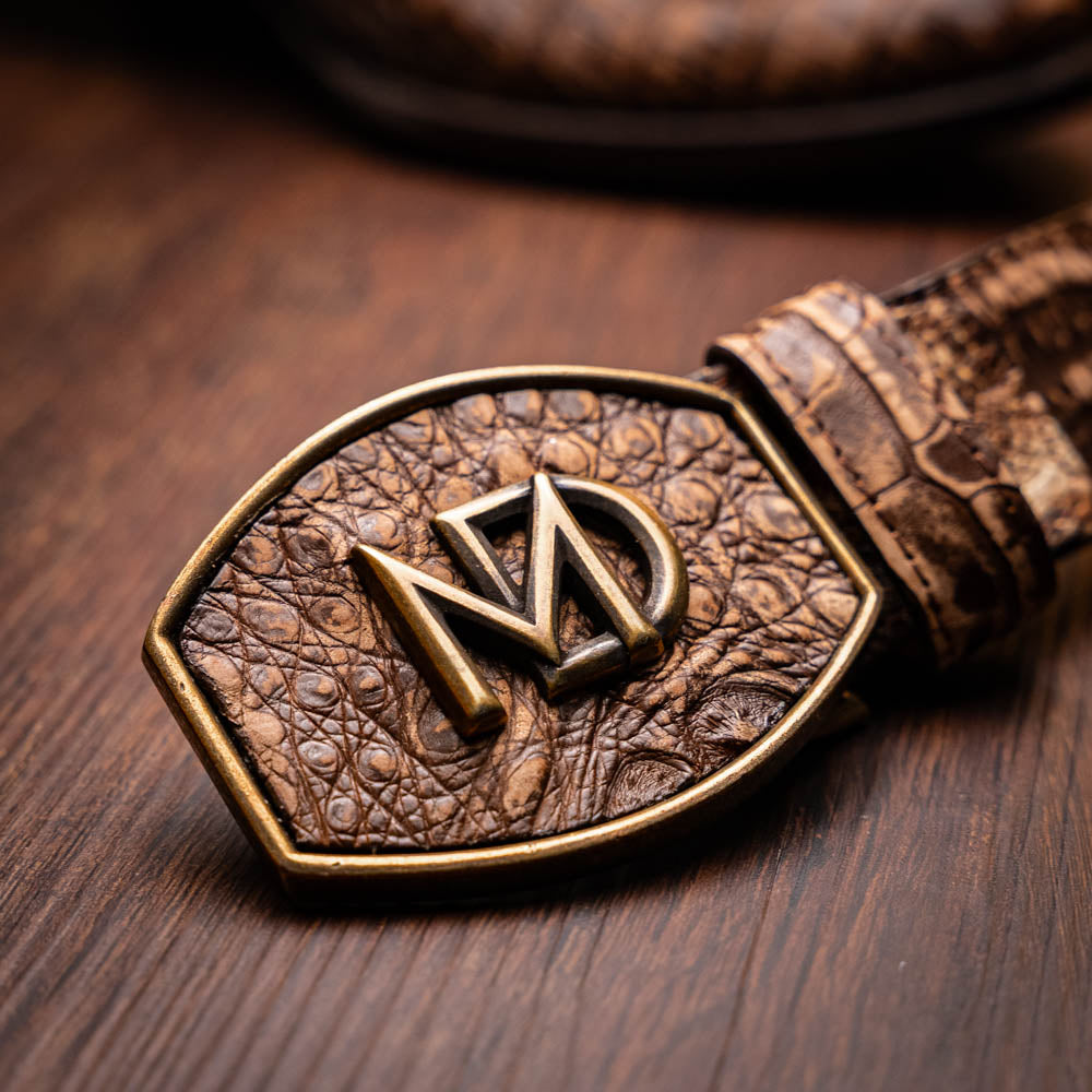 Close-up of Phoenix Stone Brown belt buckle with genuine caiman belly skin texture and metal emblem on wooden surface.
