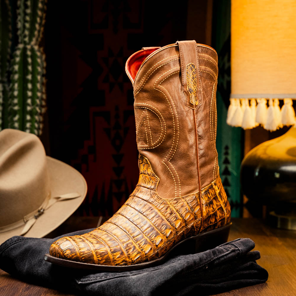Phoenix Mustard Brown cowboy boot made from genuine caiman belly skin with round toe finish, displayed on a table next to a cowboy hat.